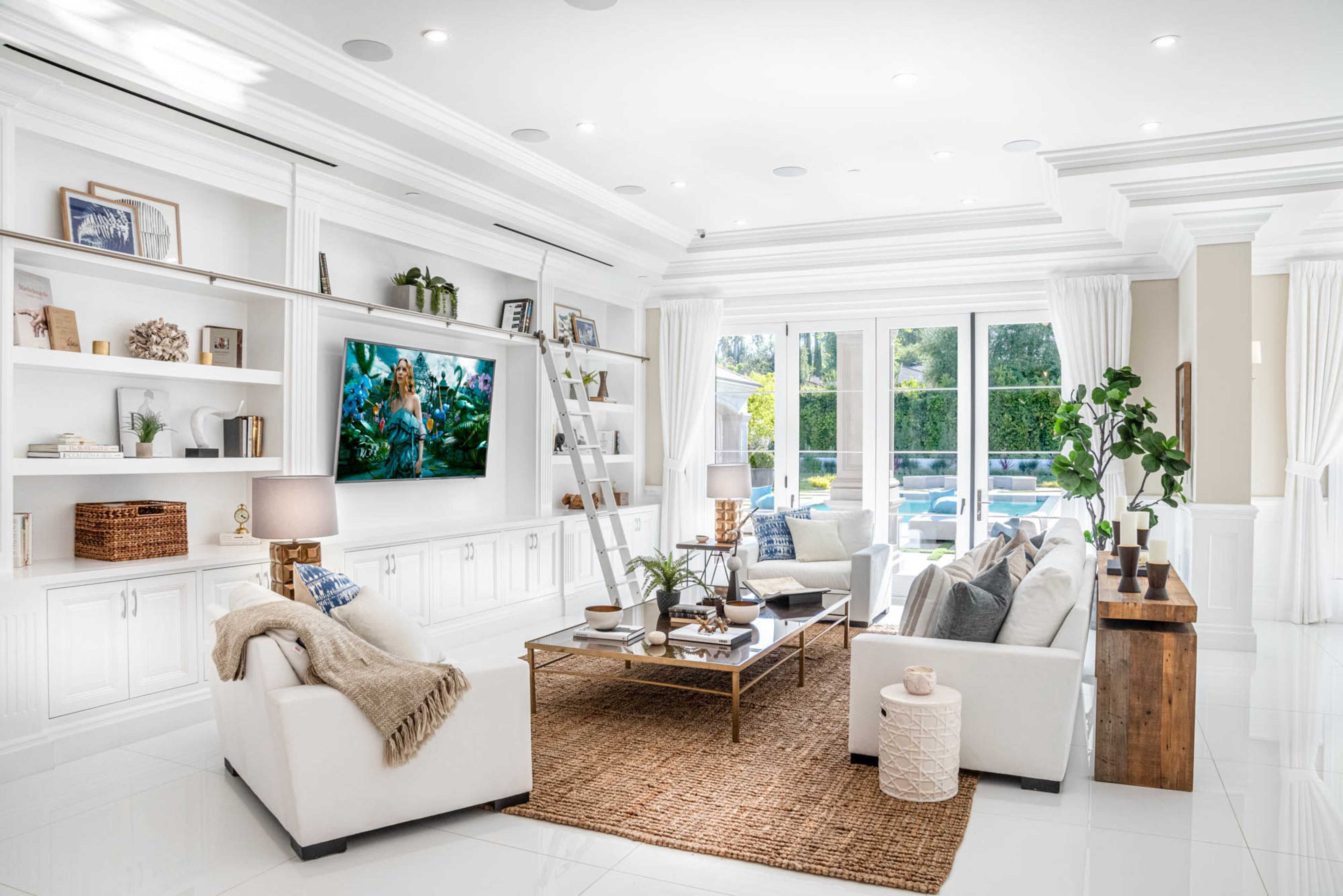 A bright, modern living room features white furniture, a large television mounted on a shelf, and large windows overlooking an outdoor space.