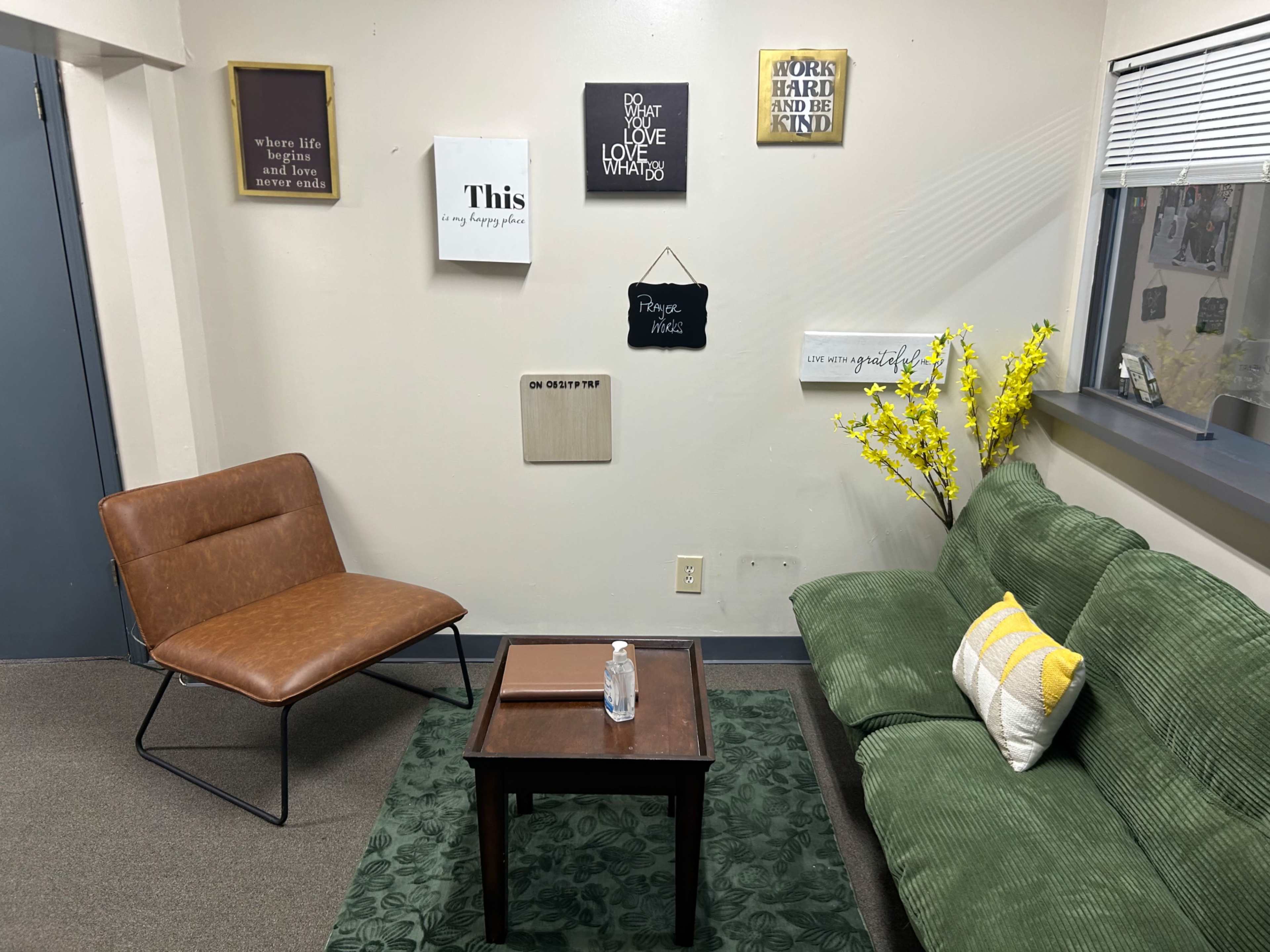 The image shows a small, softly lit room with a green couch, a brown chair, a wooden table, and various motivational wall decorations.