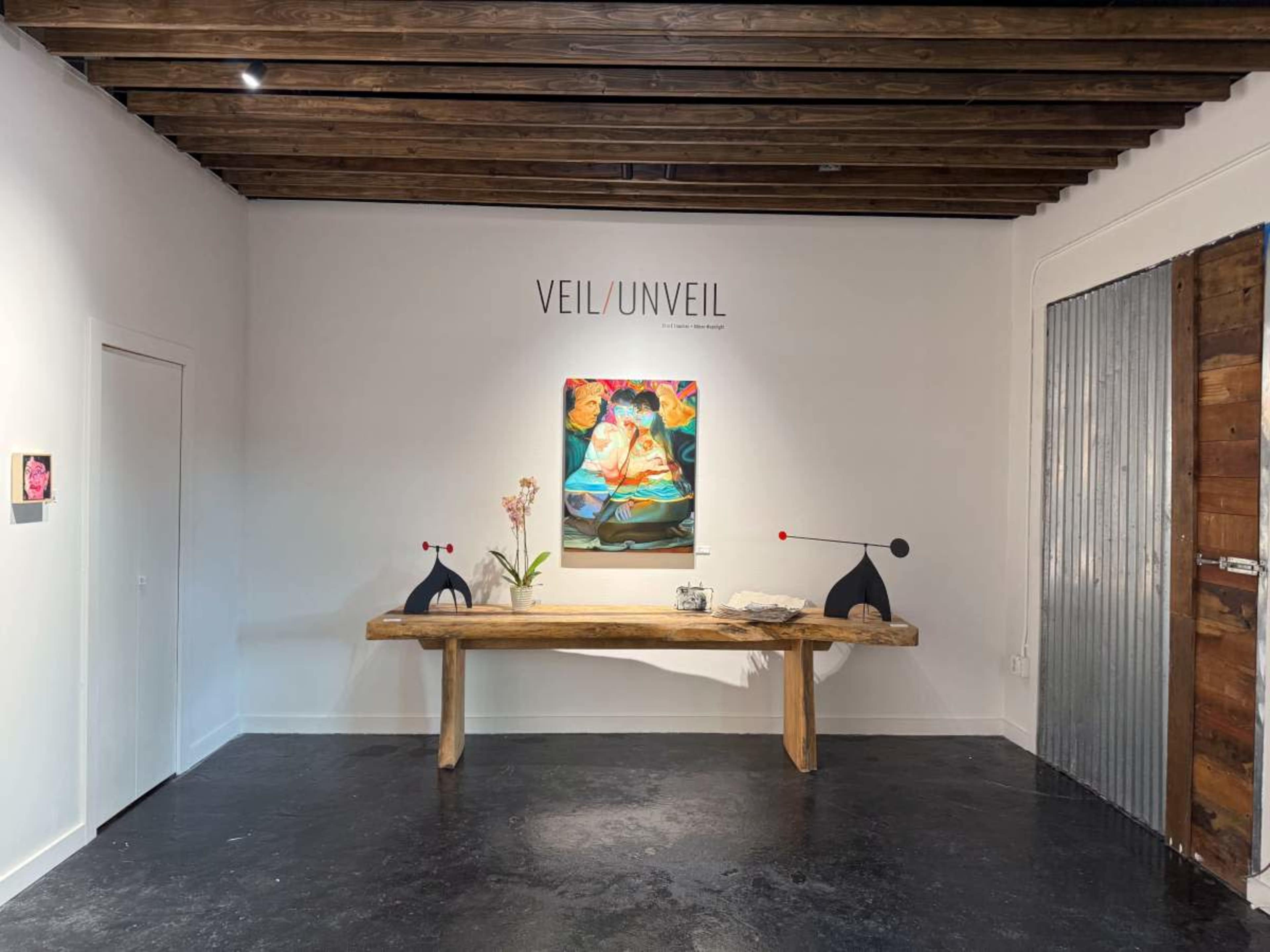 The image shows a modern art gallery space featuring a wooden table with sculptures, a painting on the wall, and minimalistic decor.