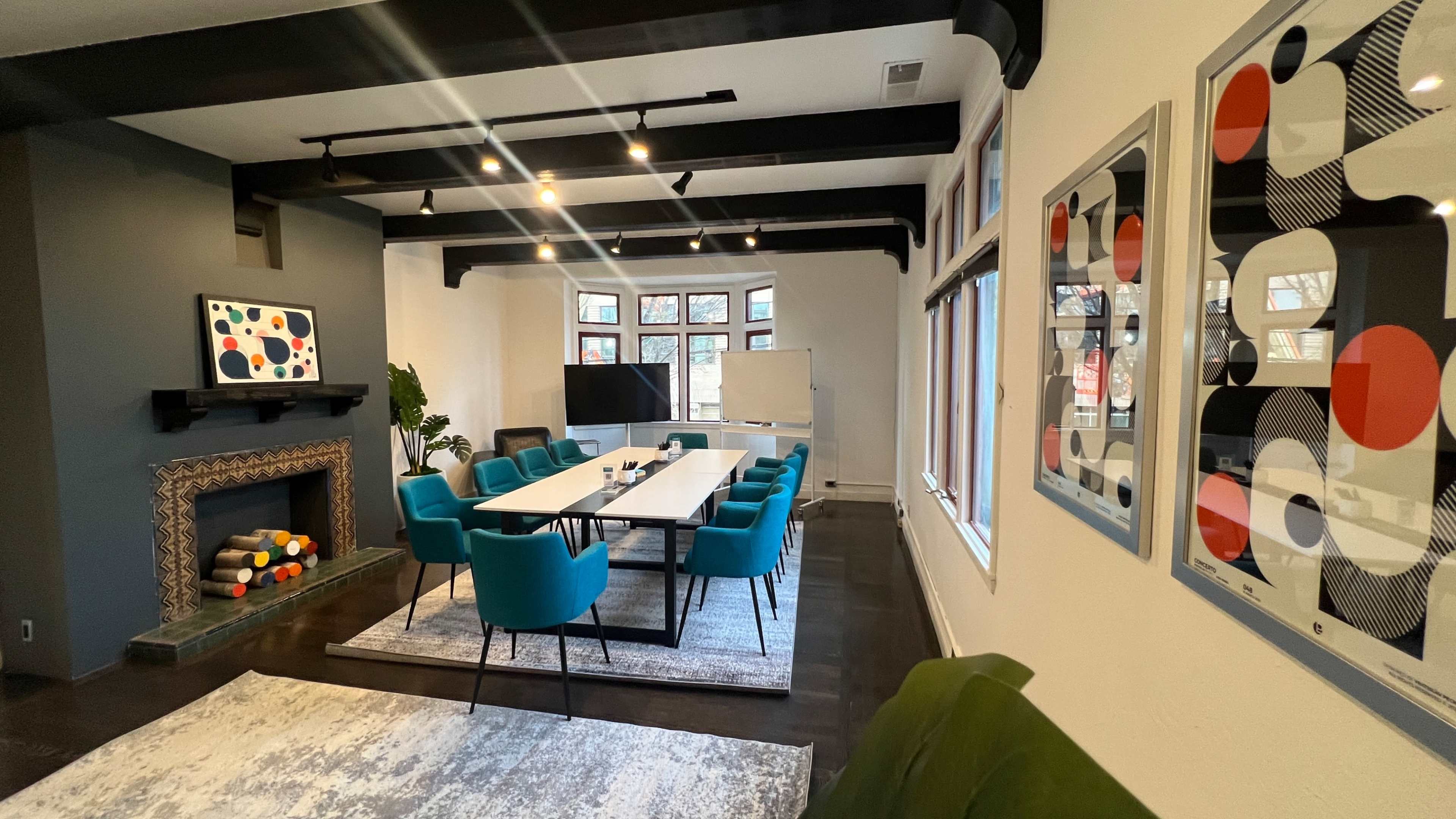 A modern conference room features a long table surrounded by teal chairs, a fireplace, and large windows with artwork on the walls.