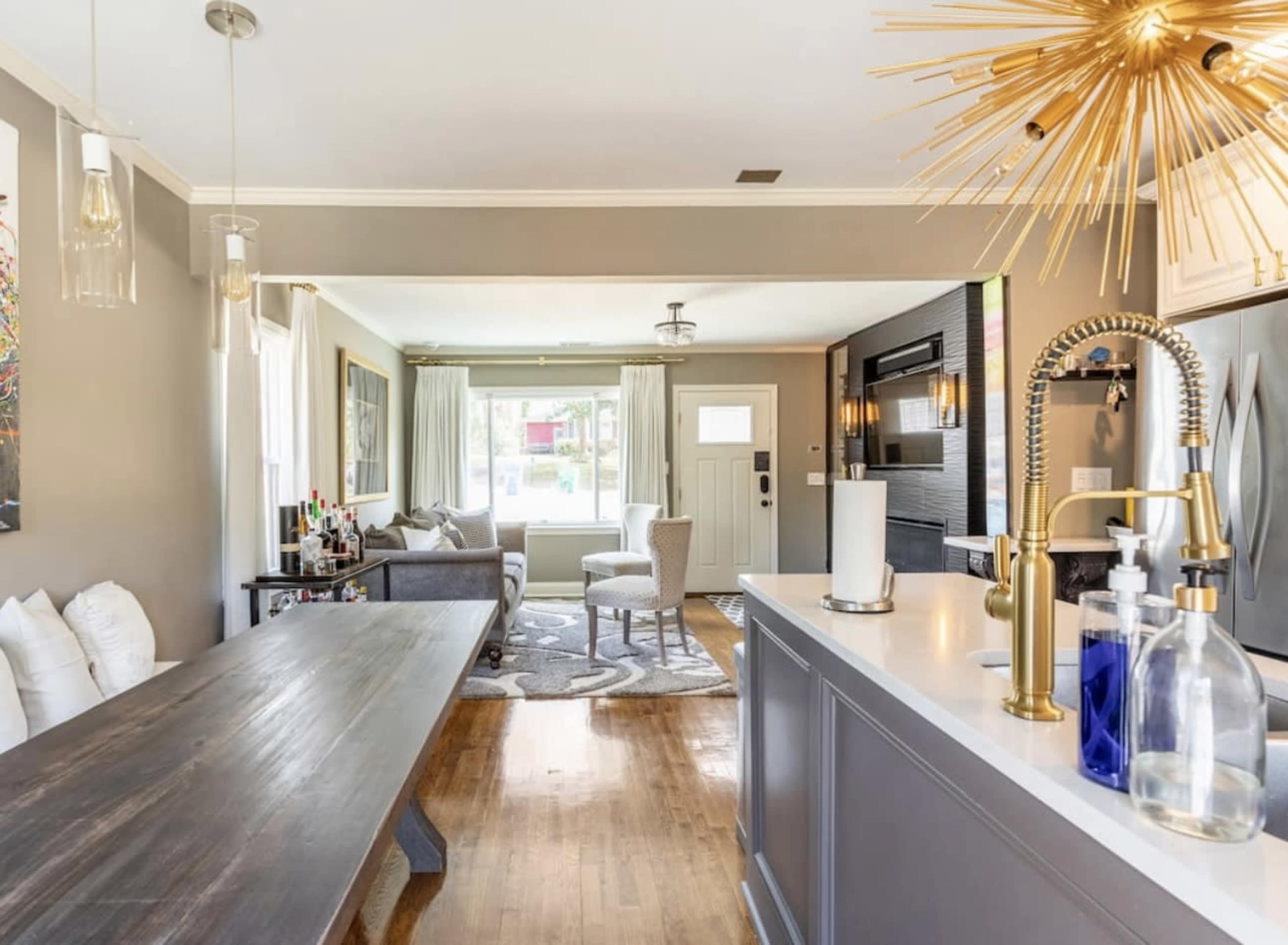 A modern kitchen and dining area with a long wooden table, stylish lighting fixtures, and a view of a living room with a sofa and large windows.