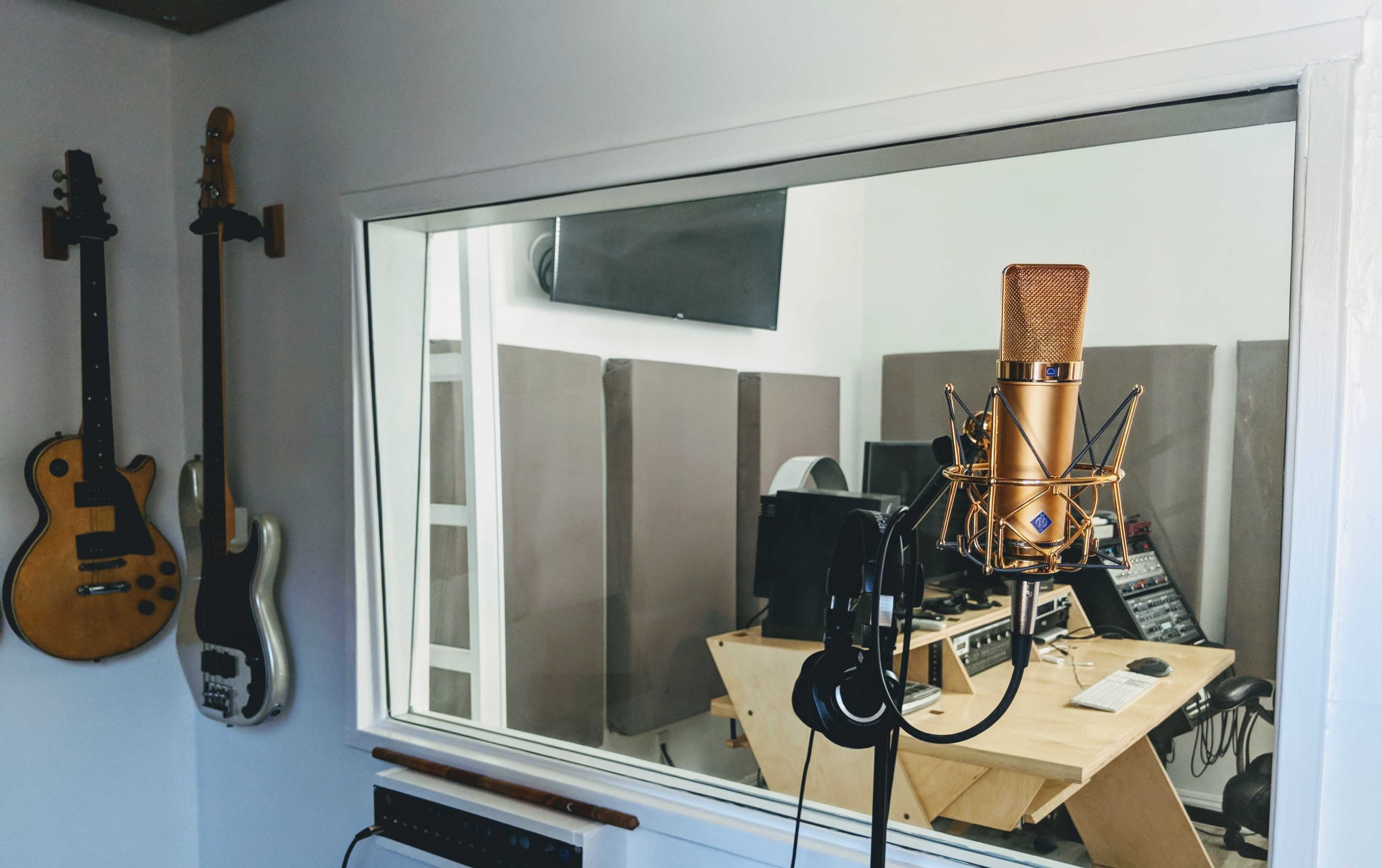 A microphone is positioned in front of a glass window leading into a music studio, which contains various instruments and equipment.