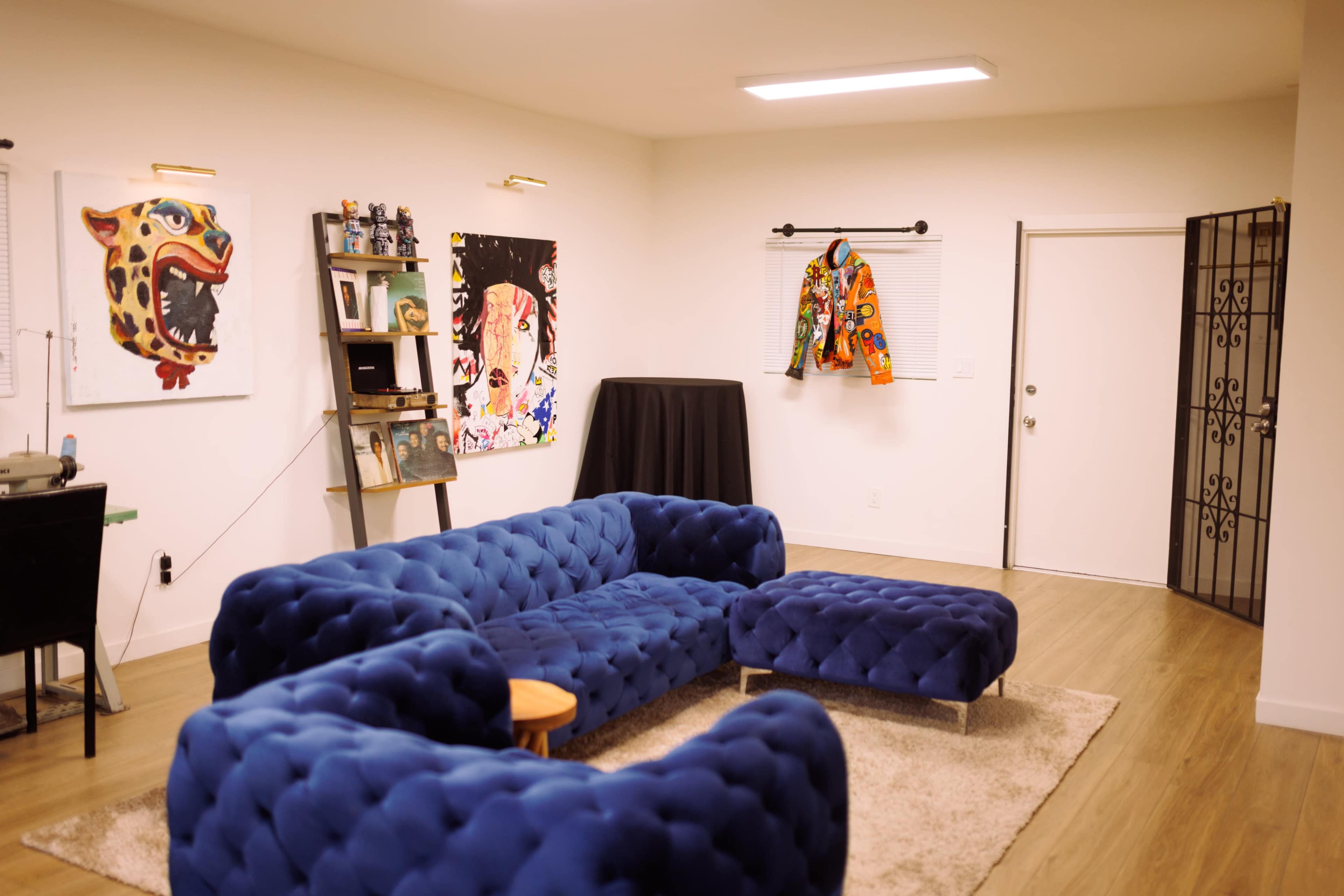 The image shows a modern, bright room featuring a blue tufted sectional sofa, a small round coffee table, artwork on the walls, and a door leading outside.