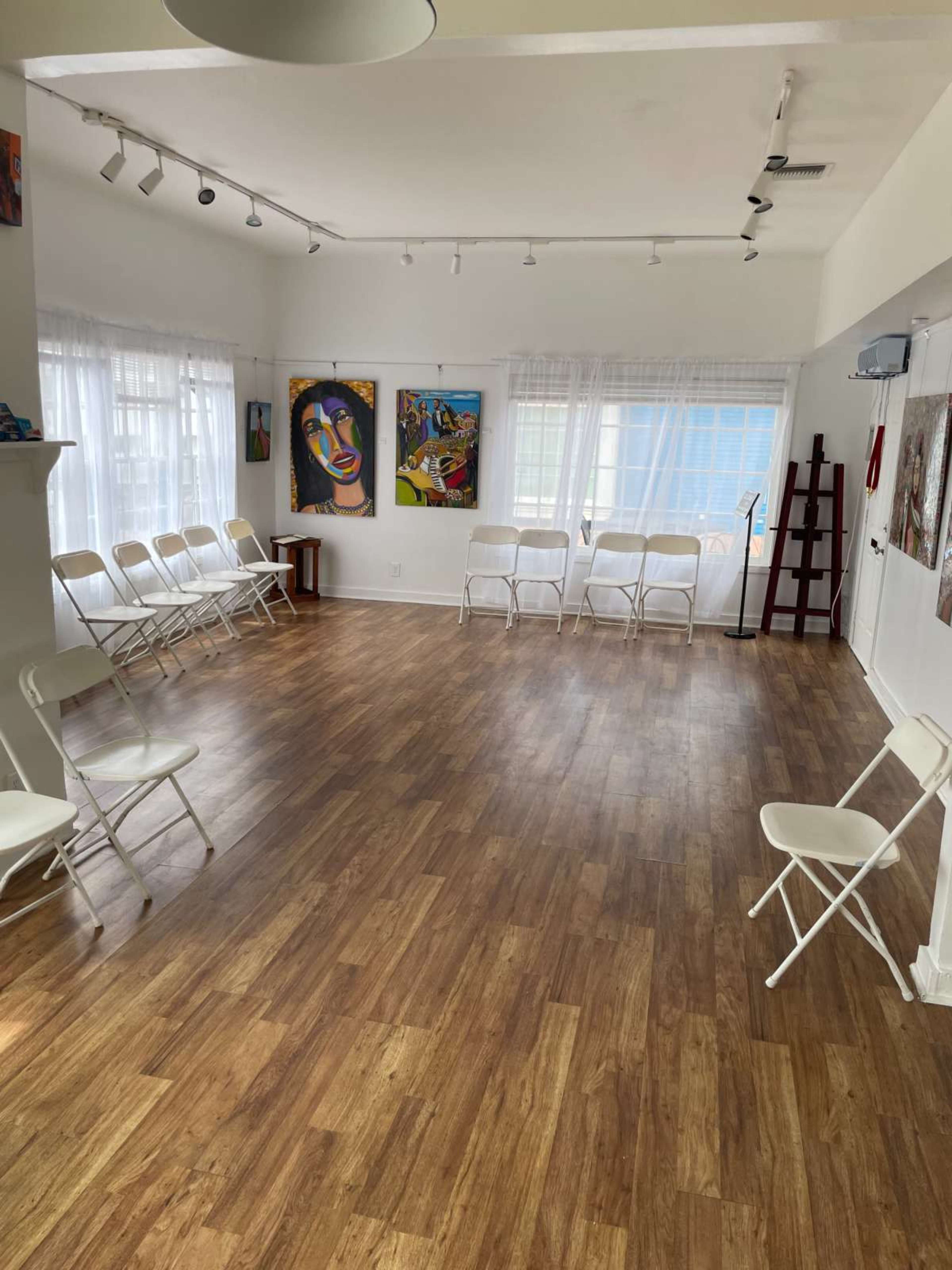 The image shows a bright, empty room with wooden flooring, several white chairs arranged in a circle, and colorful artworks on the walls.
