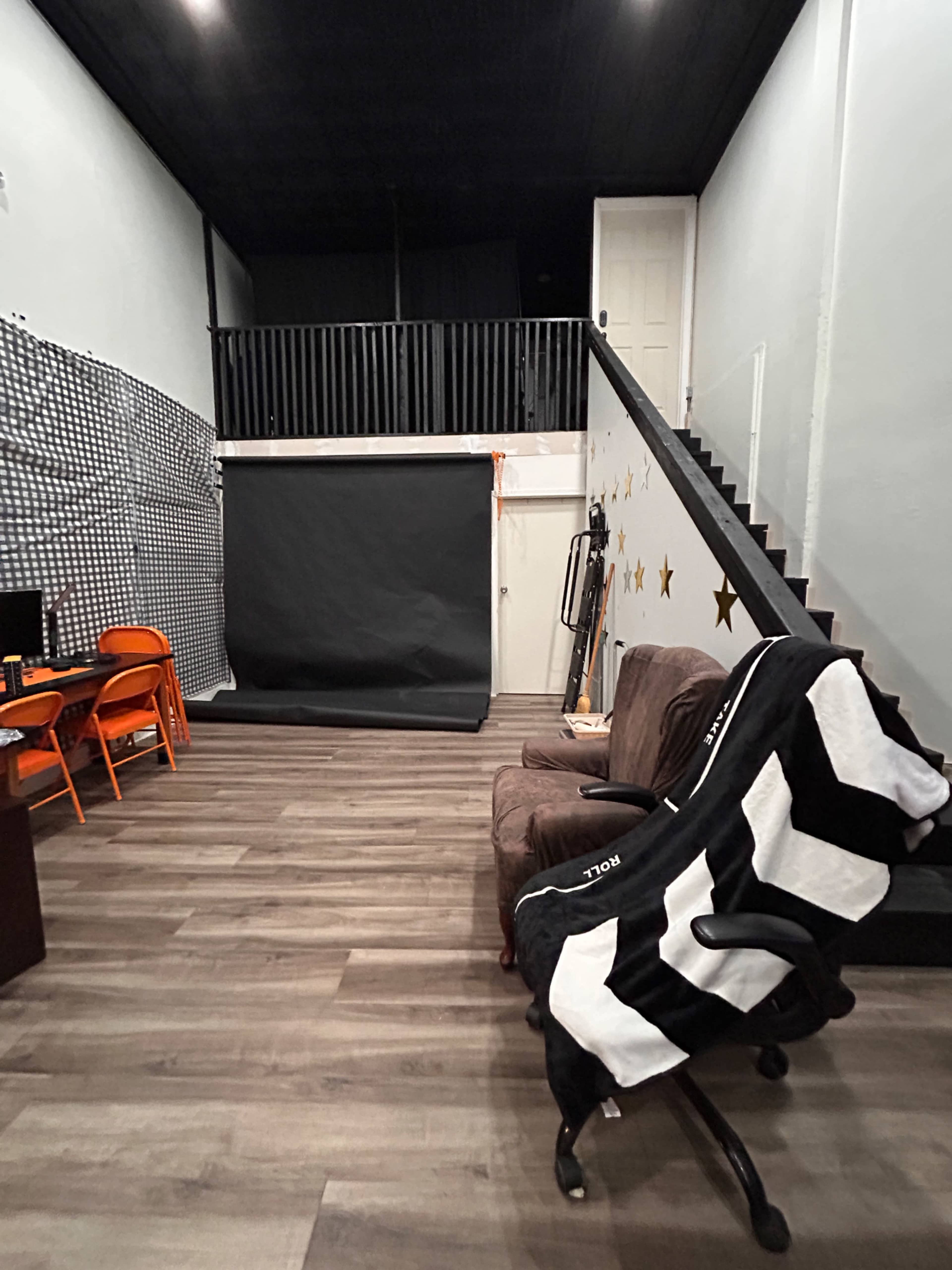 The image depicts a studio space with a dark backdrop, a staircase leading to a second level, and a cozy seating area featuring a chair and a brown couch.