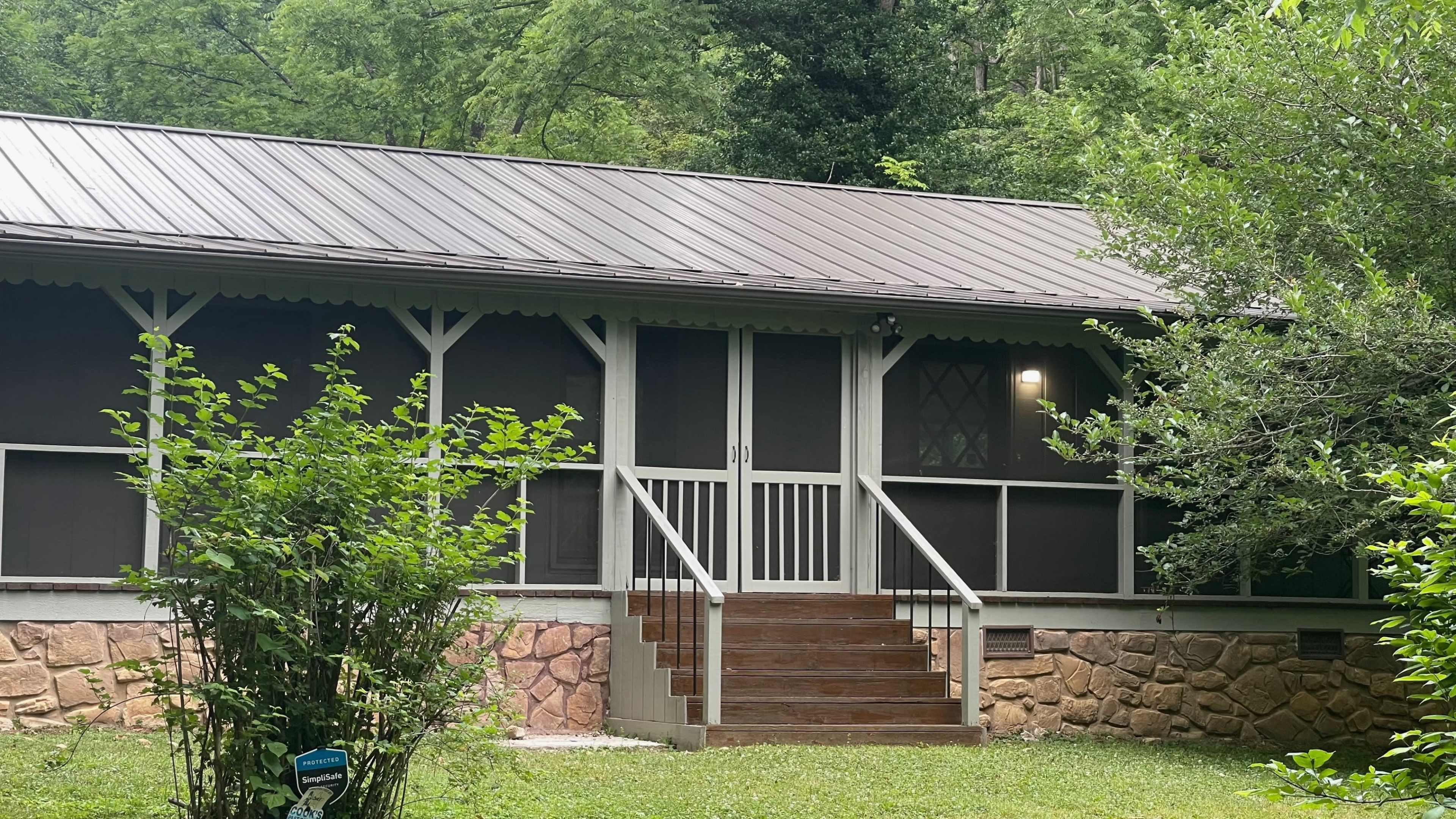 A wooden steps lead to the screened front porch of a house surrounded by greenery.