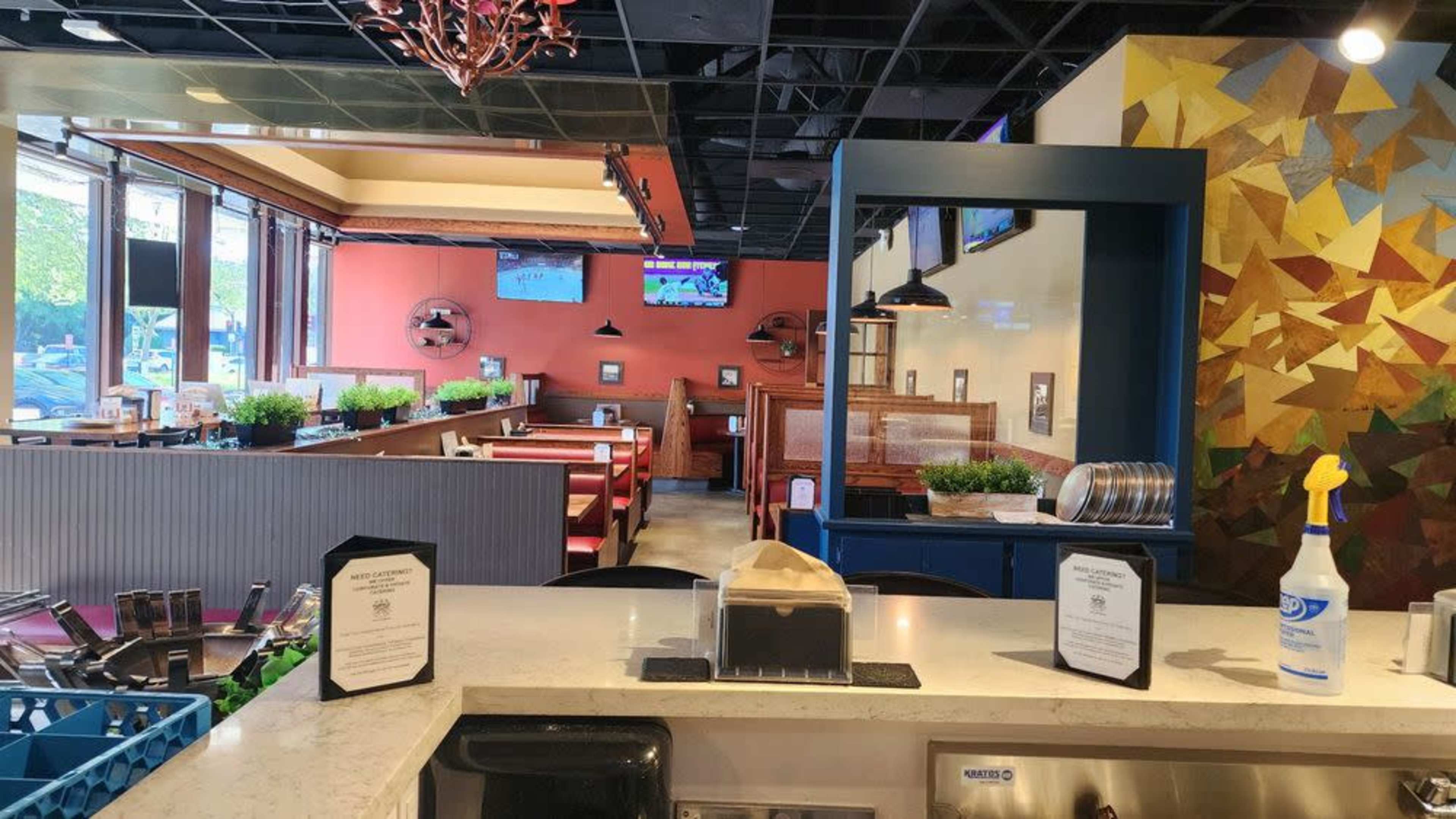 A brightly lit restaurant interior features booths and tables, with a serving counter in the foreground and television screens mounted on the walls.