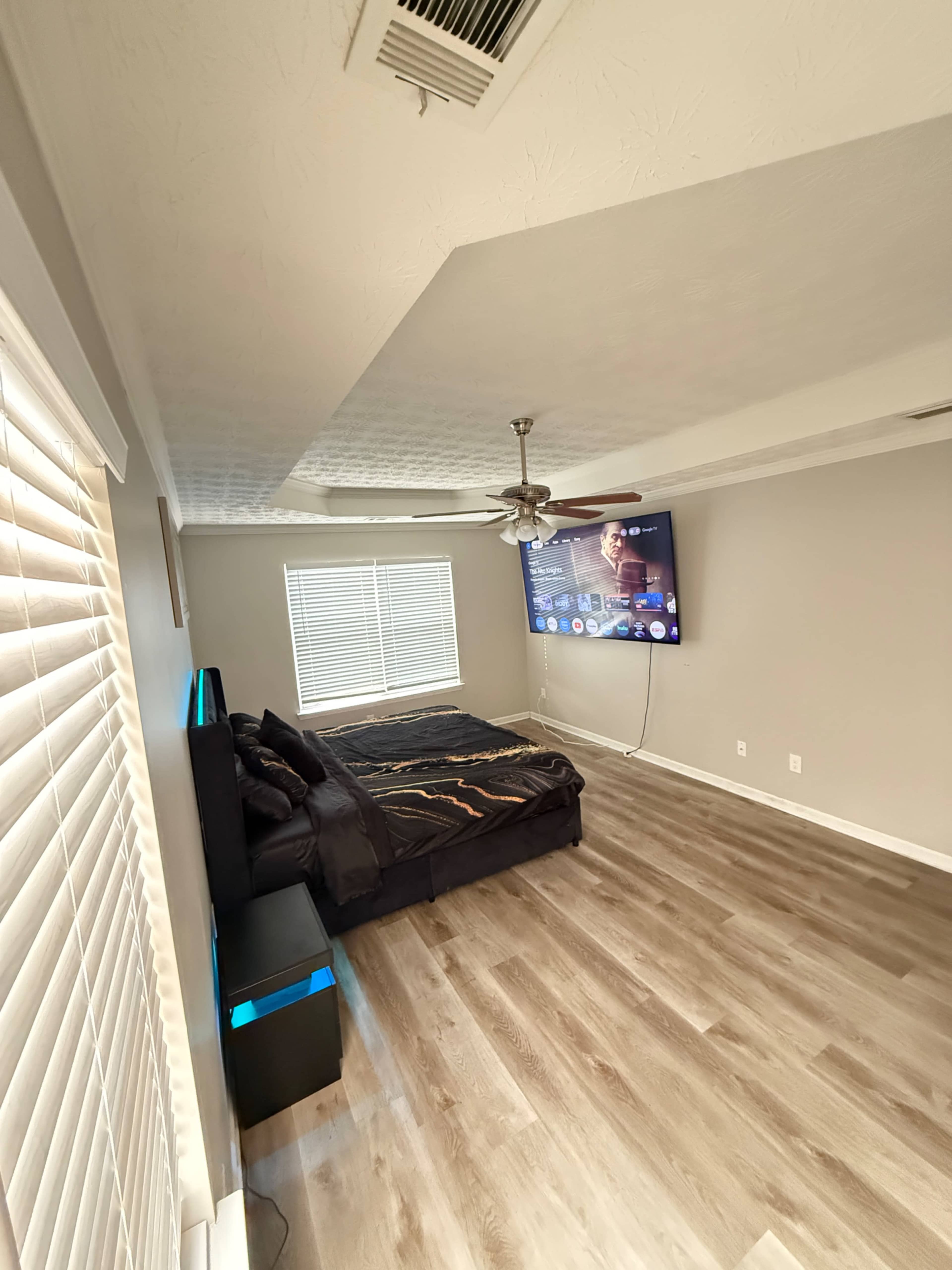 The image shows a modern bedroom with wooden flooring, a bed against the wall, a wall-mounted television, and a window with blinds.