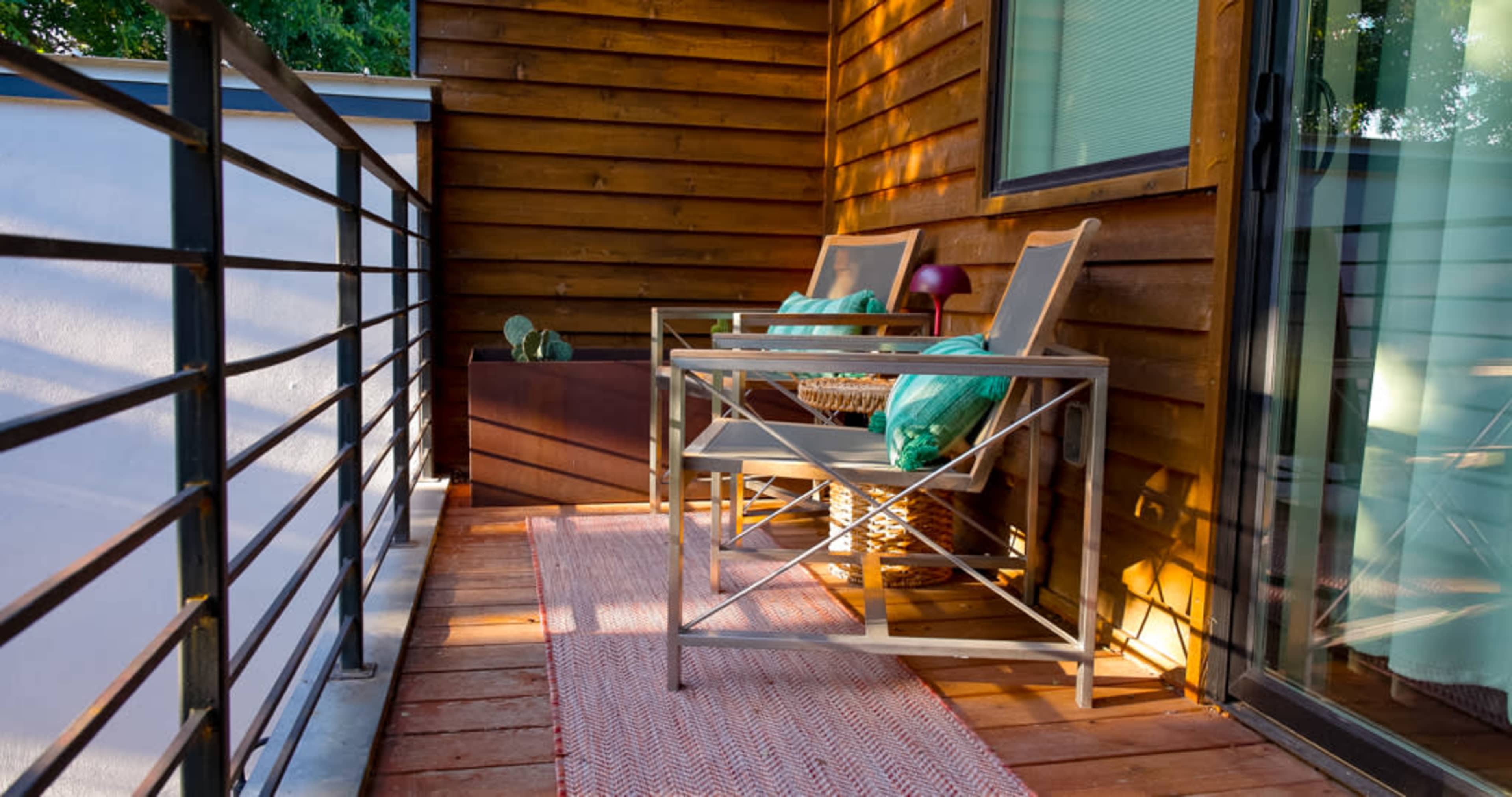 The image shows a small balcony with two metal-framed chairs, a woven basket, and a decorative rug, bordered by a wooden wall and a railing.