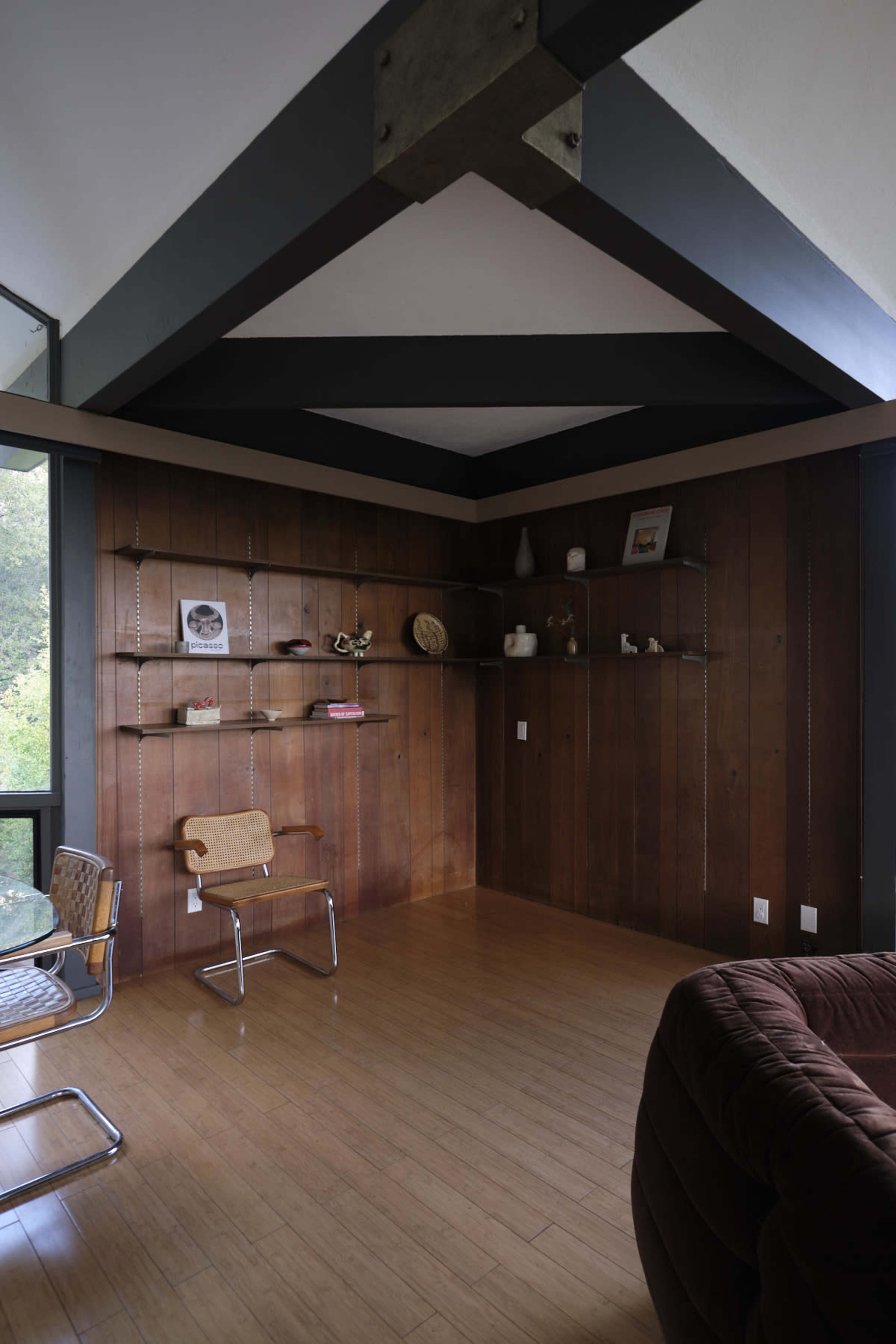 A room with wooden walls, a sloped ceiling, and minimal furniture, including a chair and a shelf displaying various decorative items.