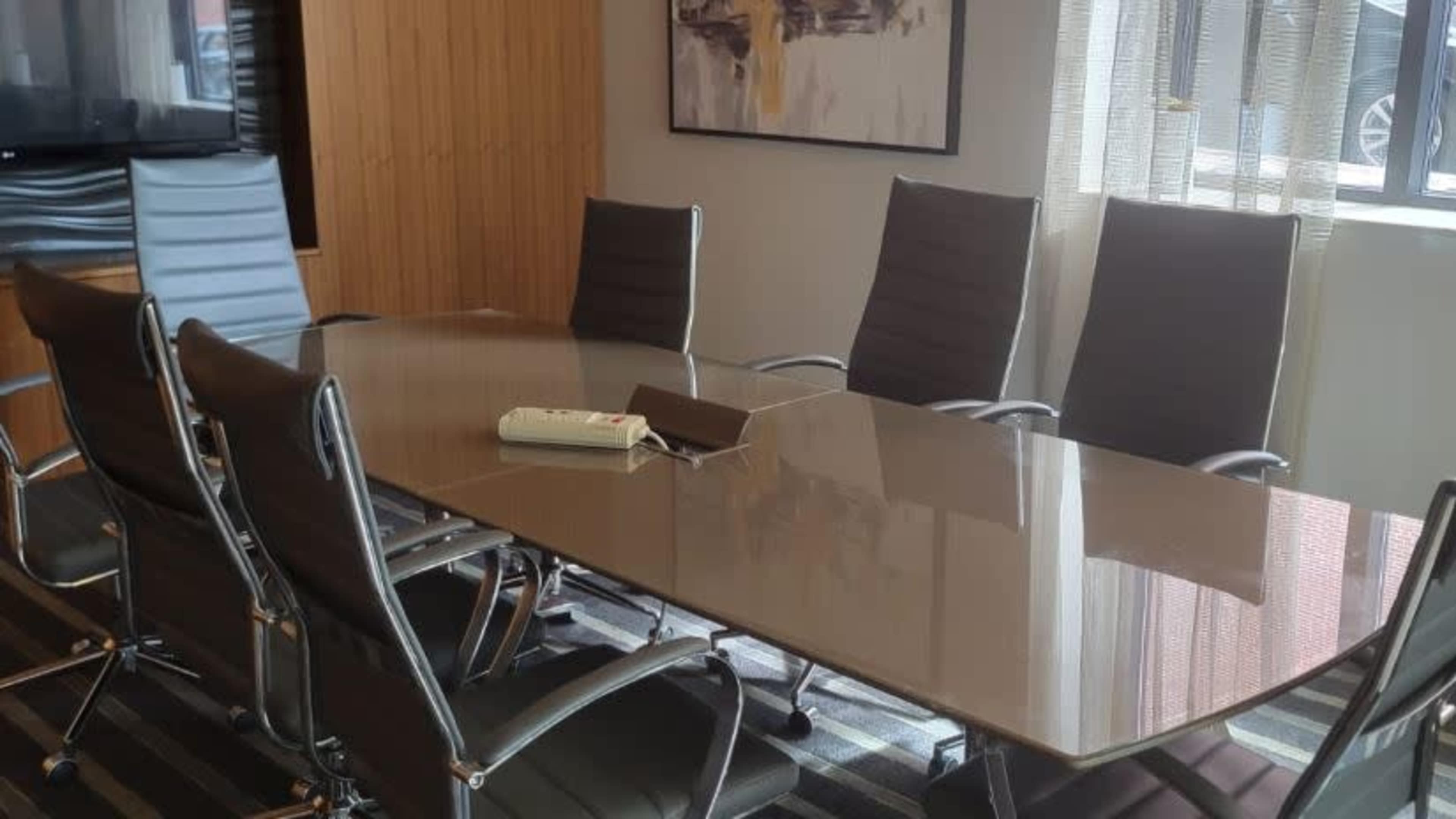 A modern conference room features a large glass table surrounded by eight sleek chairs, with natural light streaming through a nearby window.