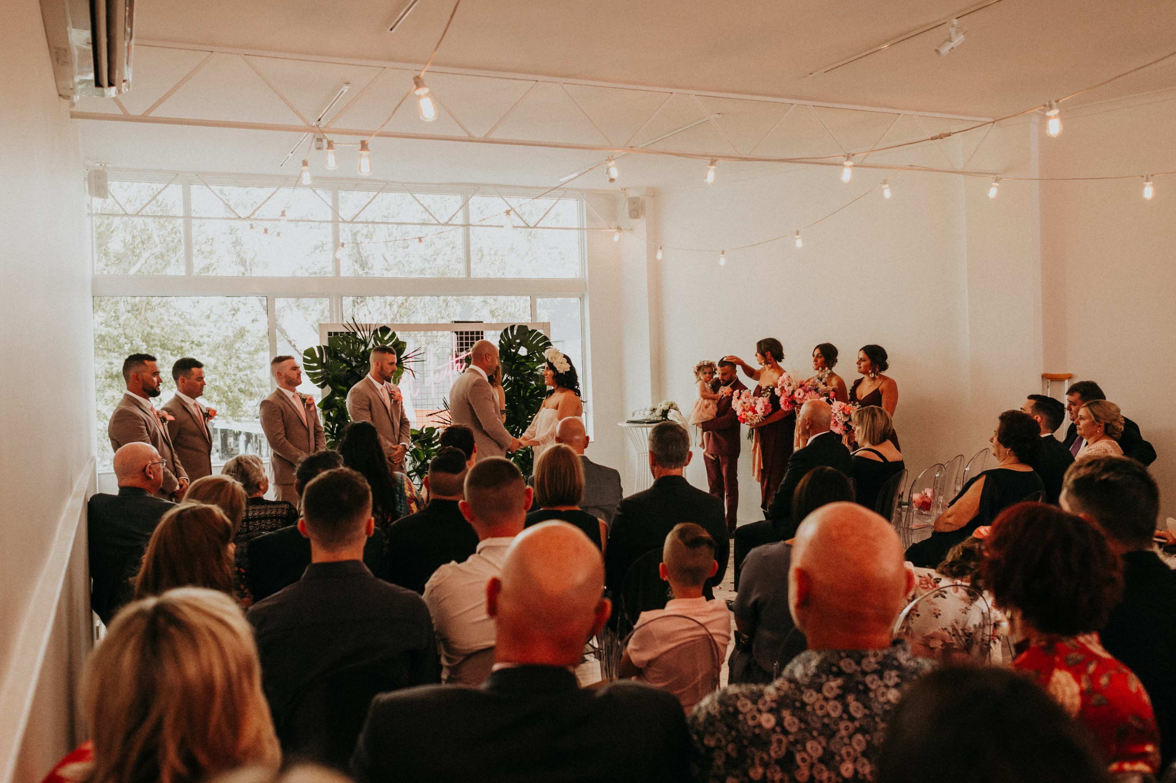A wedding ceremony is taking place in a well-lit indoor venue with guests seated in rows, facing the couple and officiant at the front.