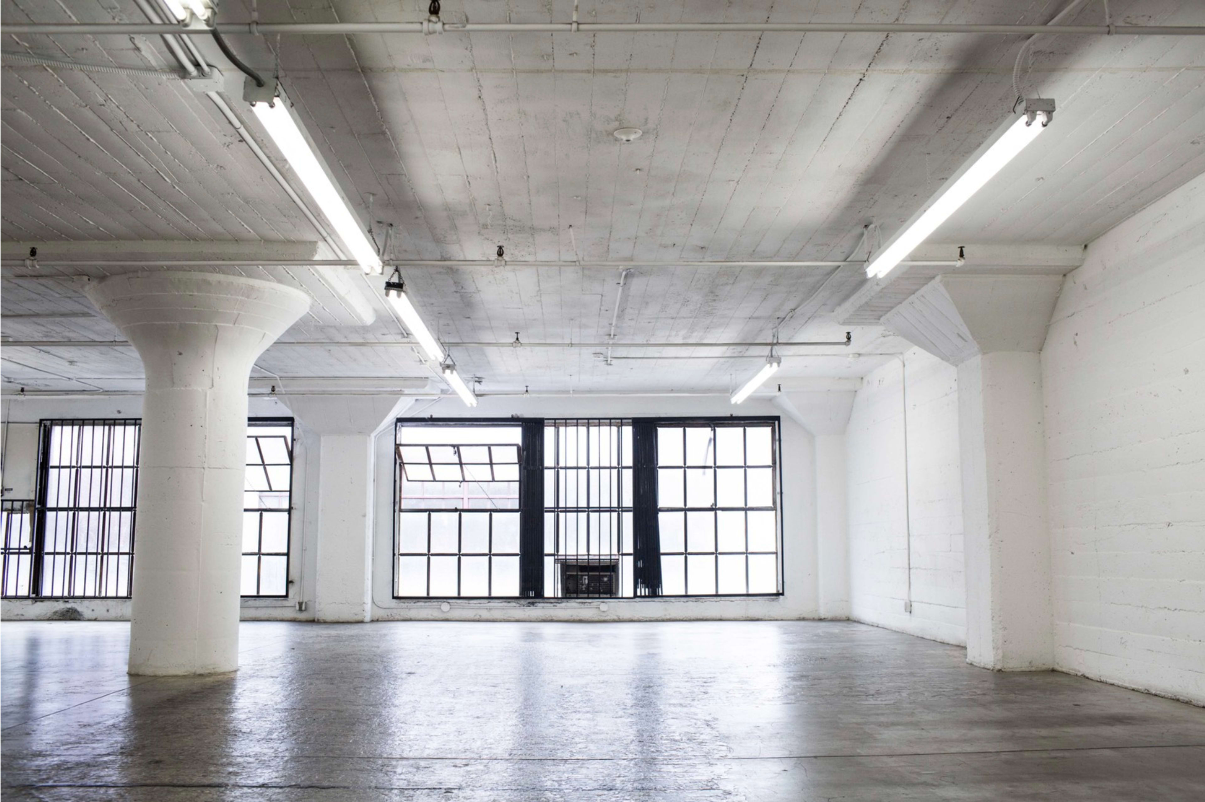 A spacious, empty room with concrete walls, high ceilings, and large windows allowing natural light to enter.
