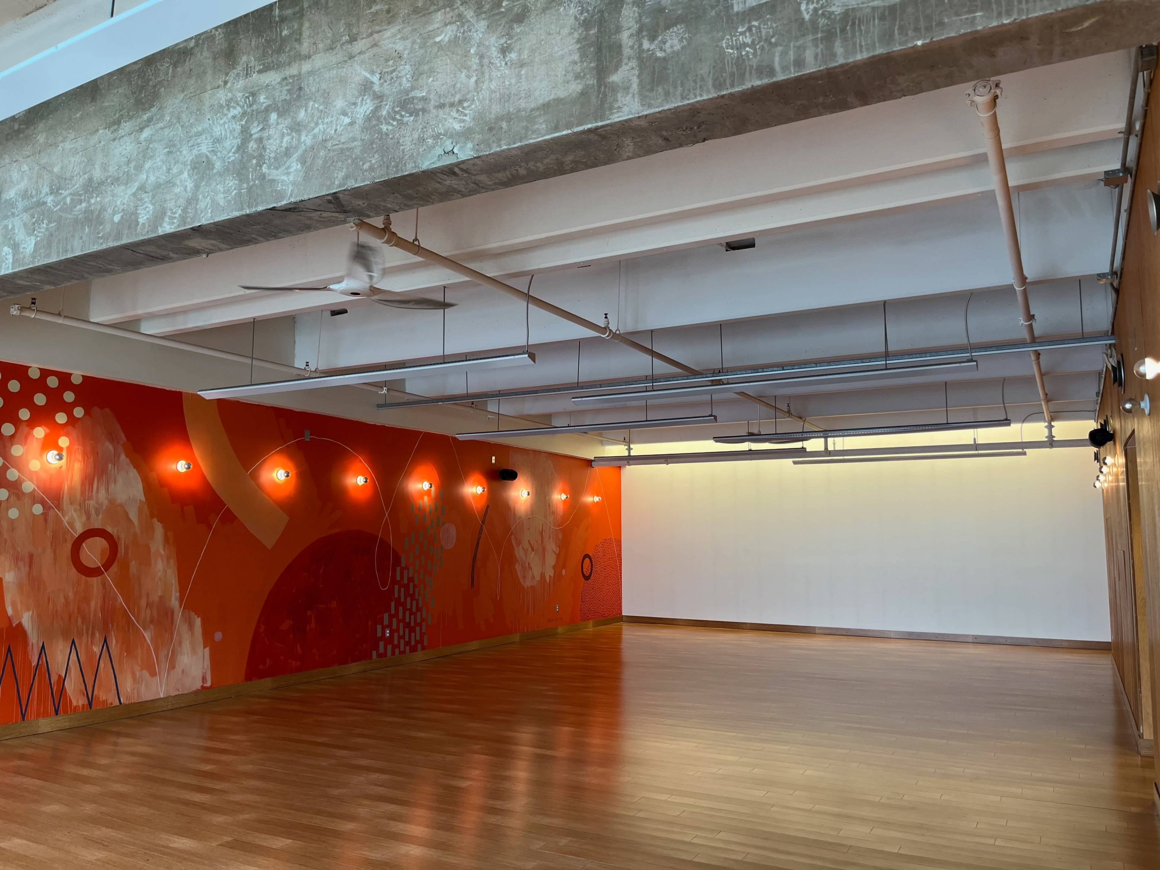 A spacious room with wooden flooring and a brightly colored mural on one wall, along with overhead lights and ceiling fans.