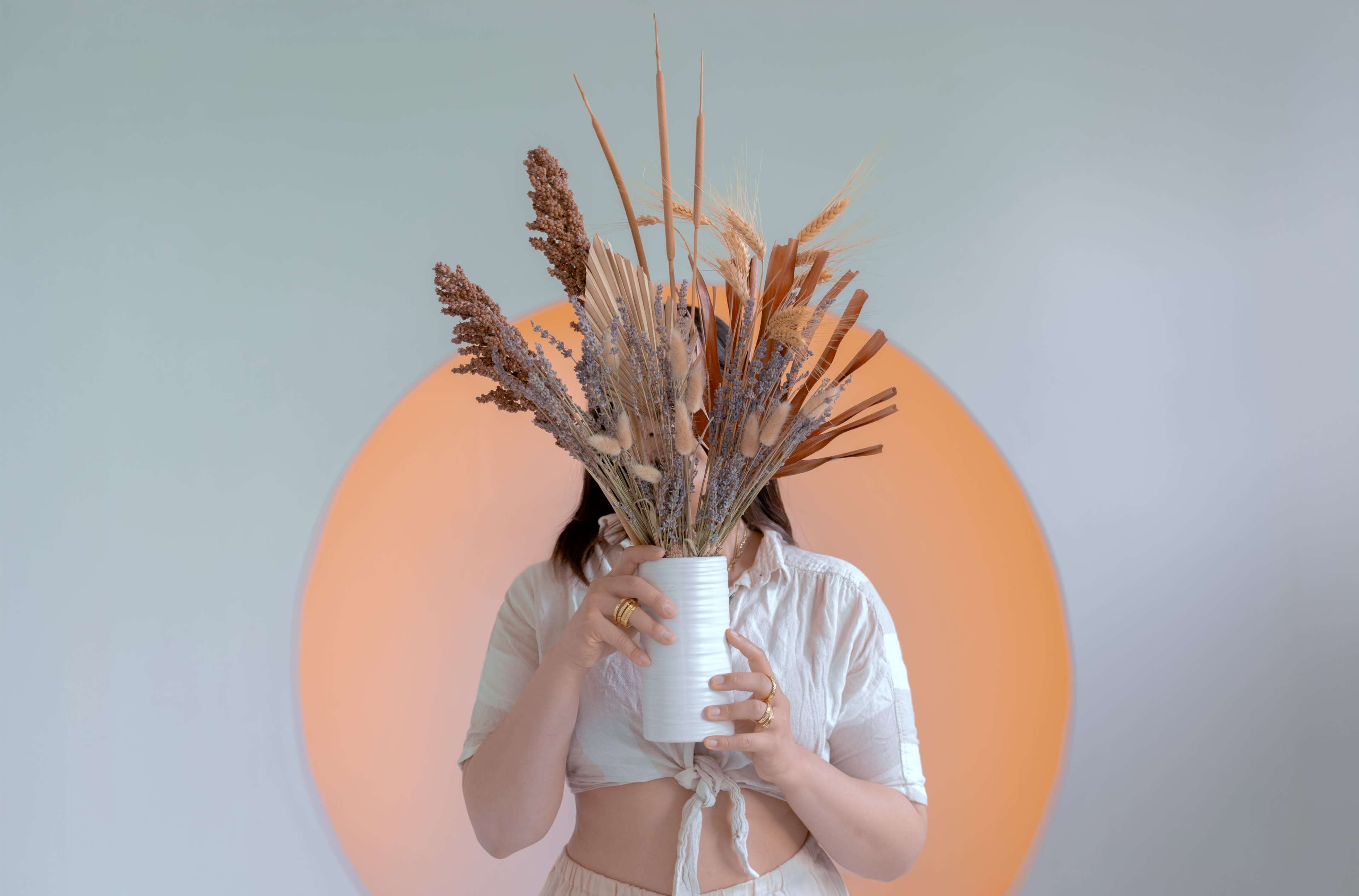 A person holds a white vase filled with dried flowers in front of their face against a backdrop of a circular orange design.
