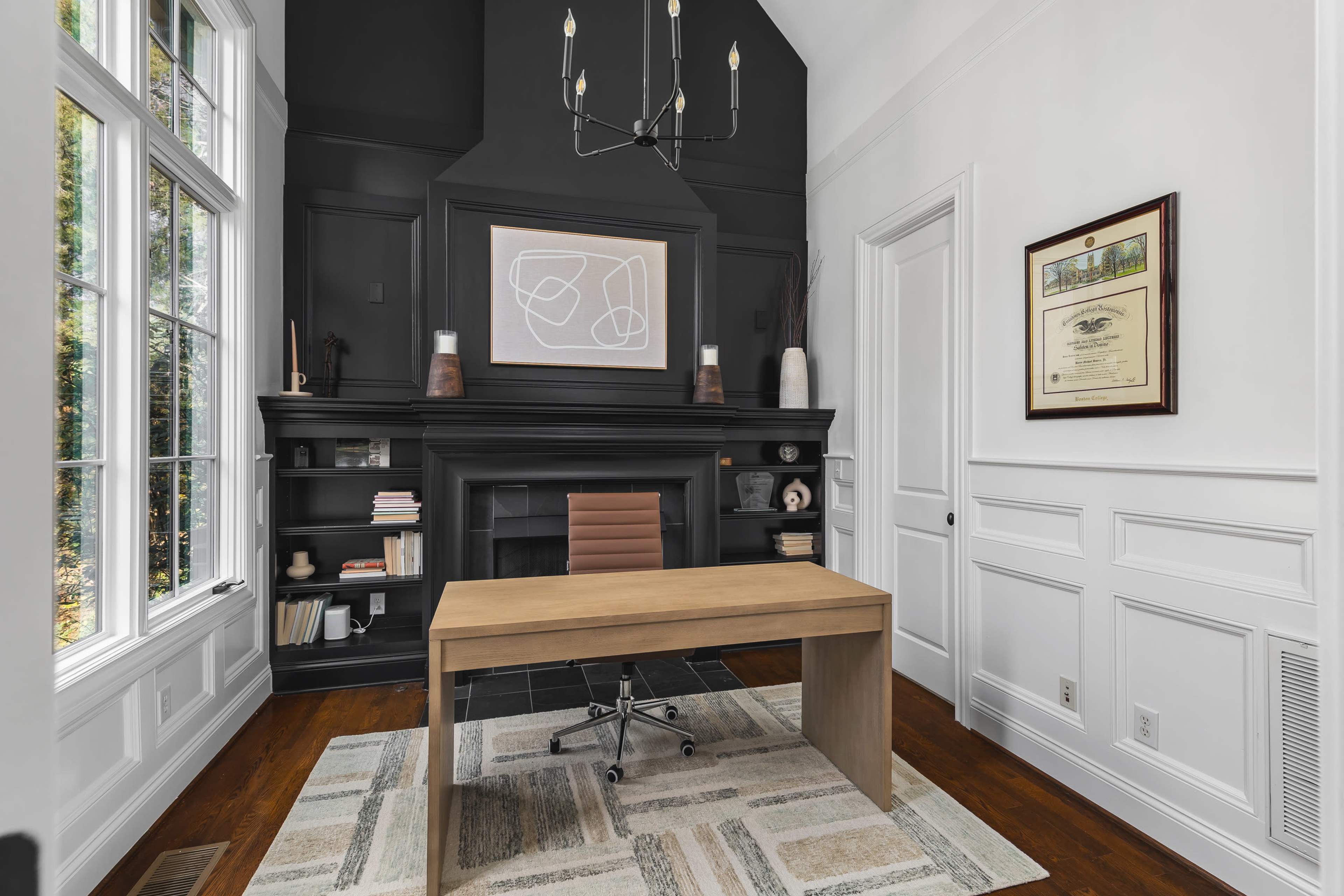 A modern office with a wooden desk facing a large black fireplace, surrounded by white walls and tall windows that let in natural light.