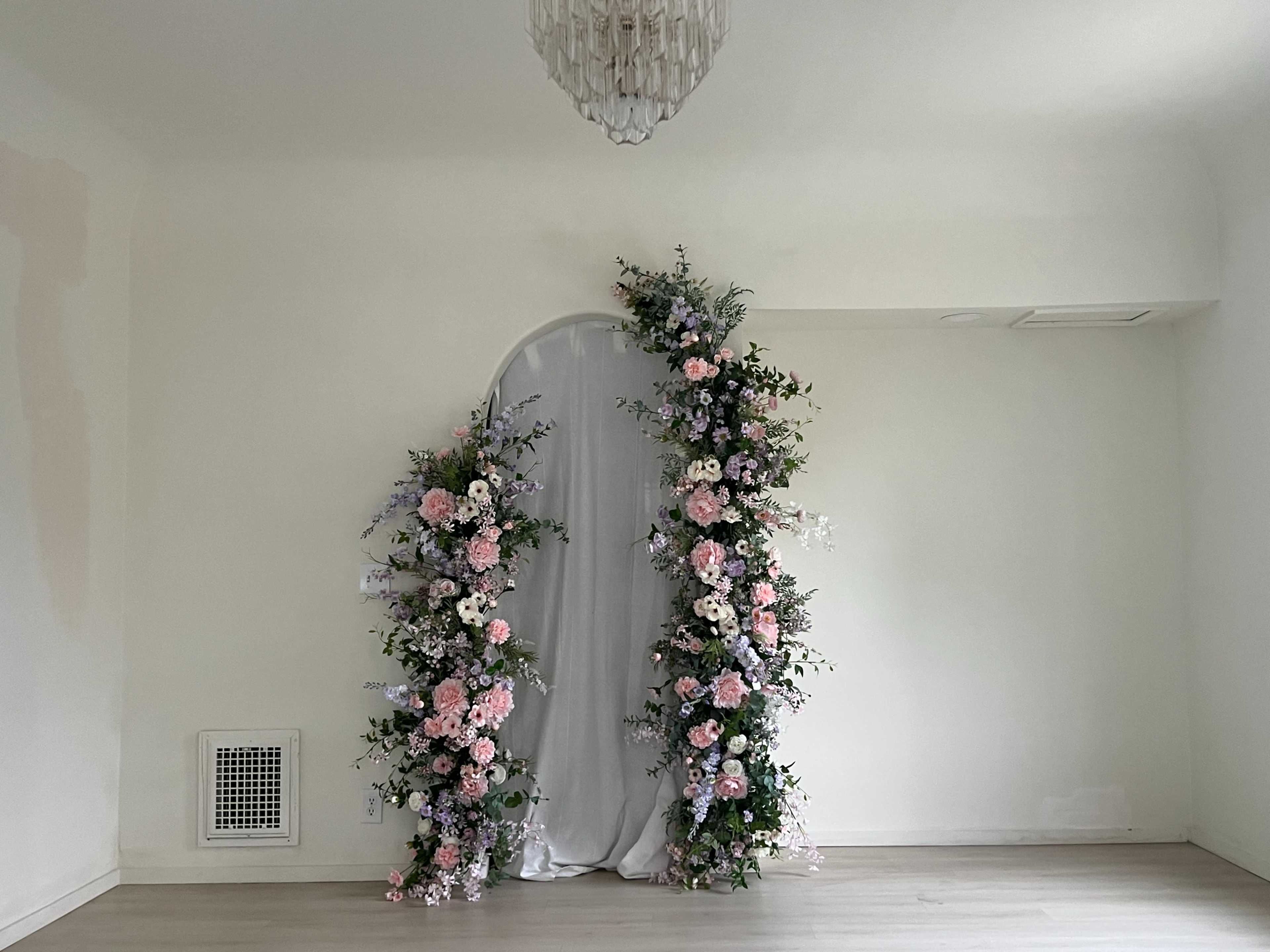 The image shows a floral archway adorned with pink and white flowers on either side of a gray fabric backdrop in a room with light-colored walls and wooden flooring.
