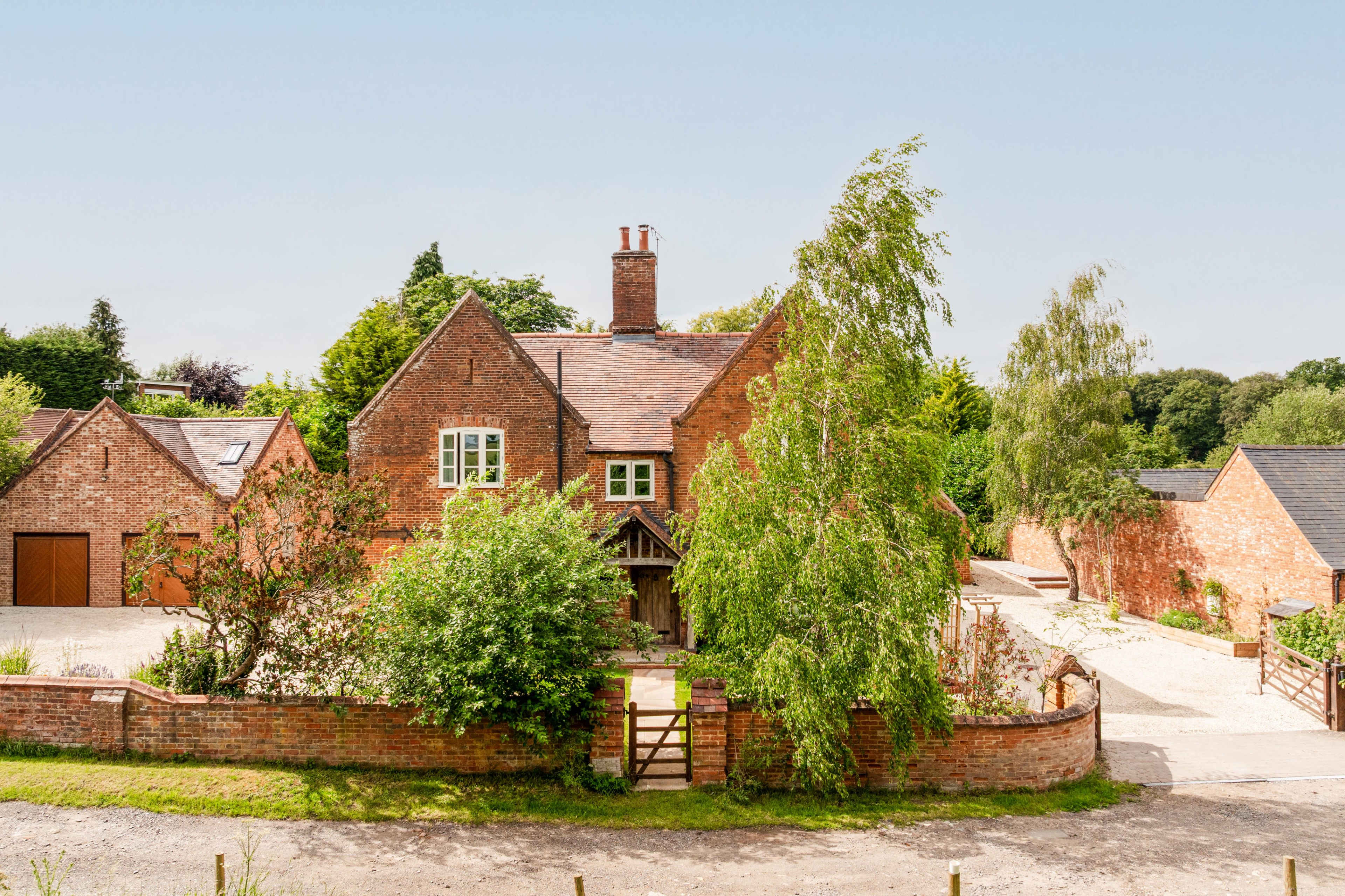 A red-brick house with a sloping roof is surrounded by greenery and a gravel driveway.