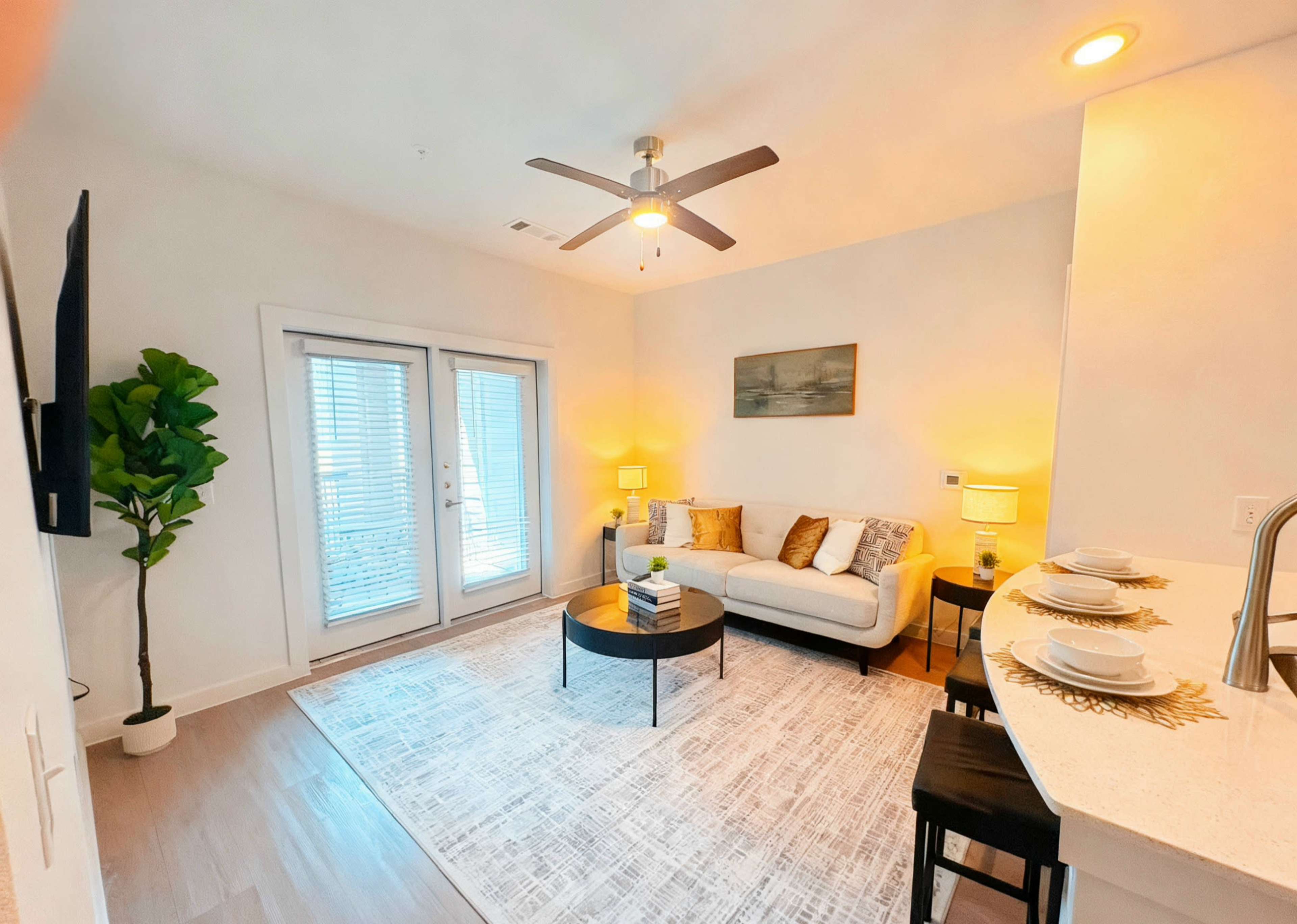 The image shows a modern living room with a light-colored sofa, a round coffee table, and a large area rug, featuring sliding glass doors that lead to a balcony.