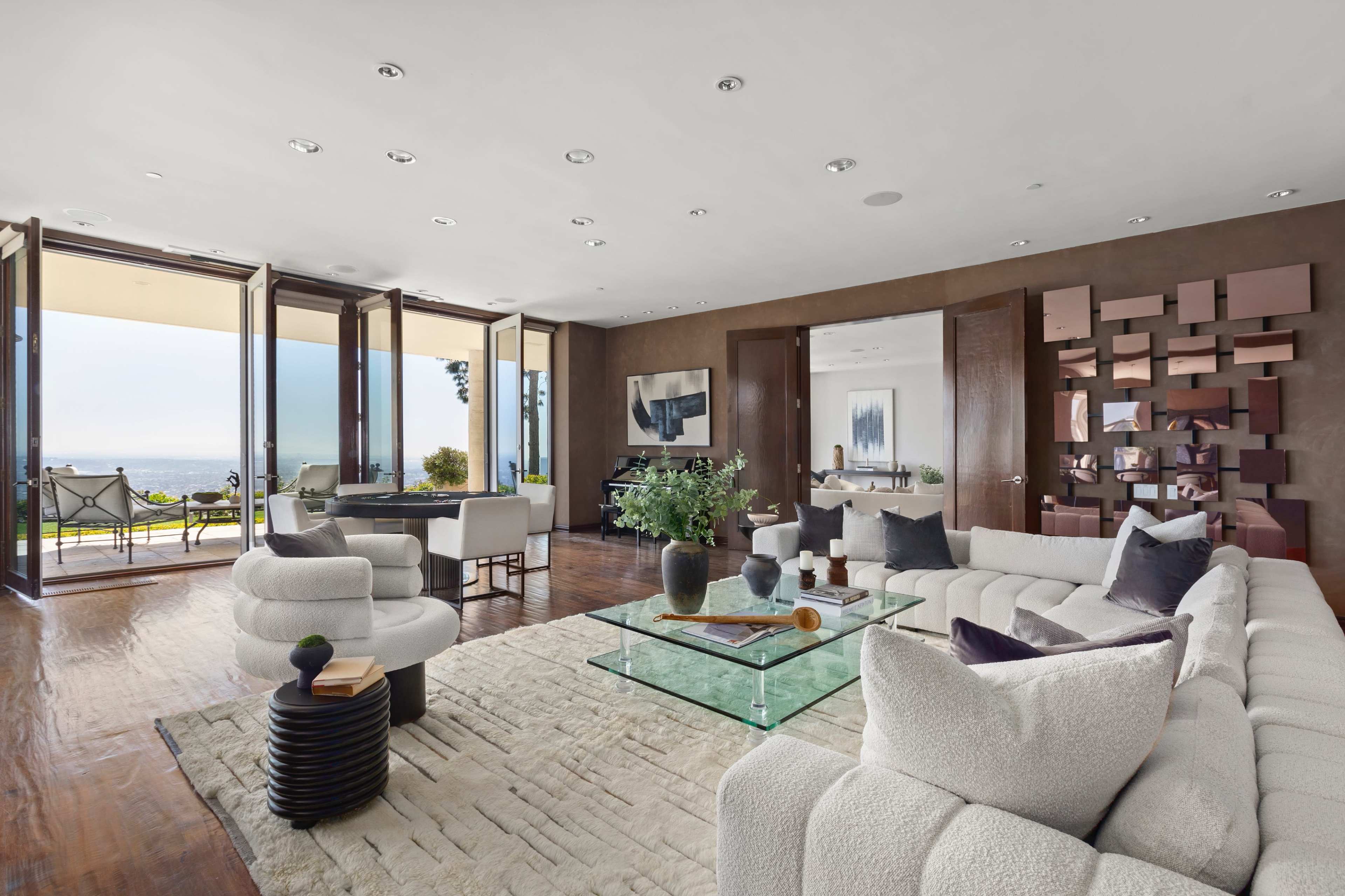 The image depicts a modern living room with large windows offering a view of the exterior, featuring minimalist furniture and a neutral color palette.