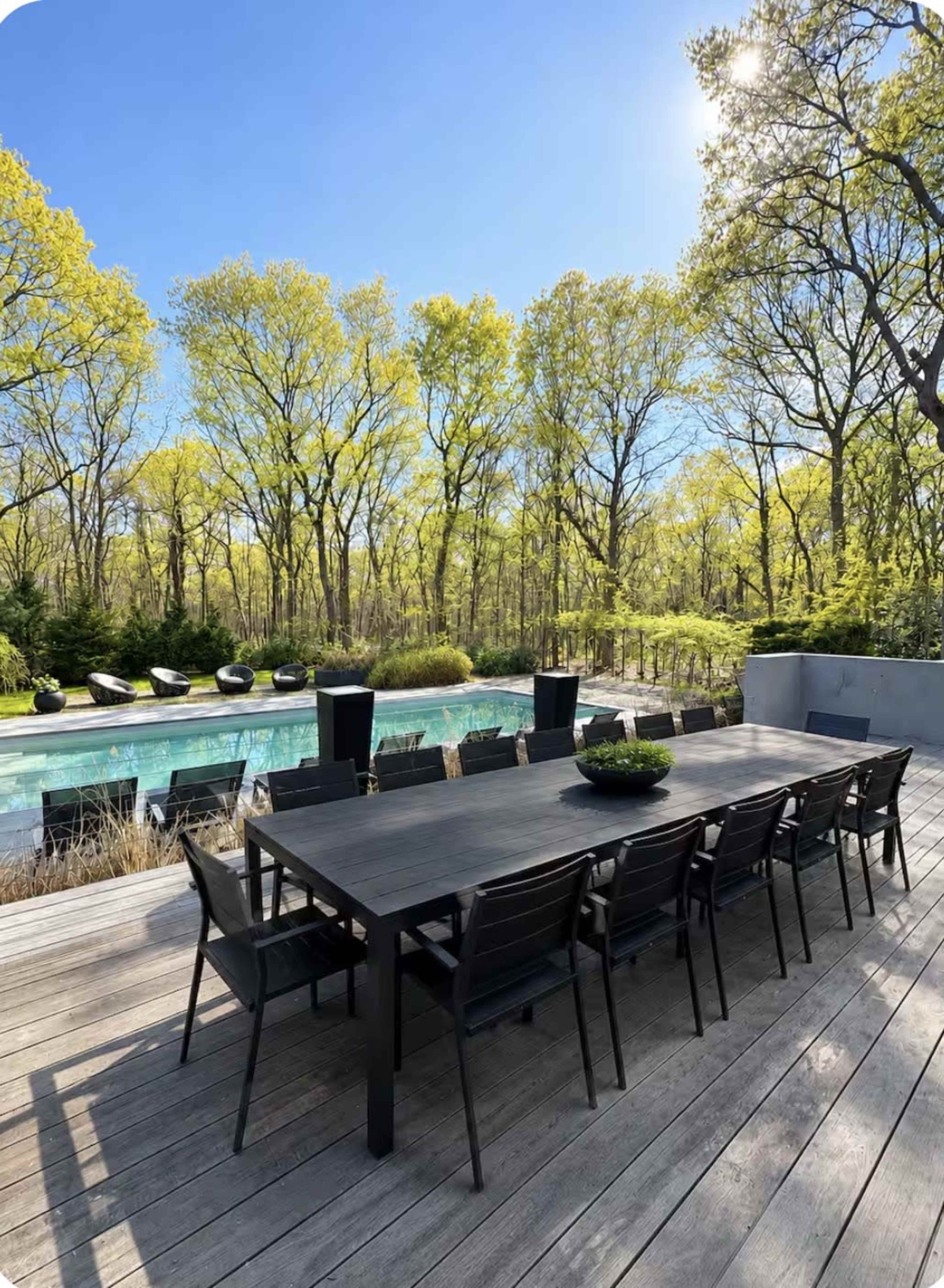 A large wooden dining table with black chairs is set beside a swimming pool surrounded by trees with fresh green leaves.