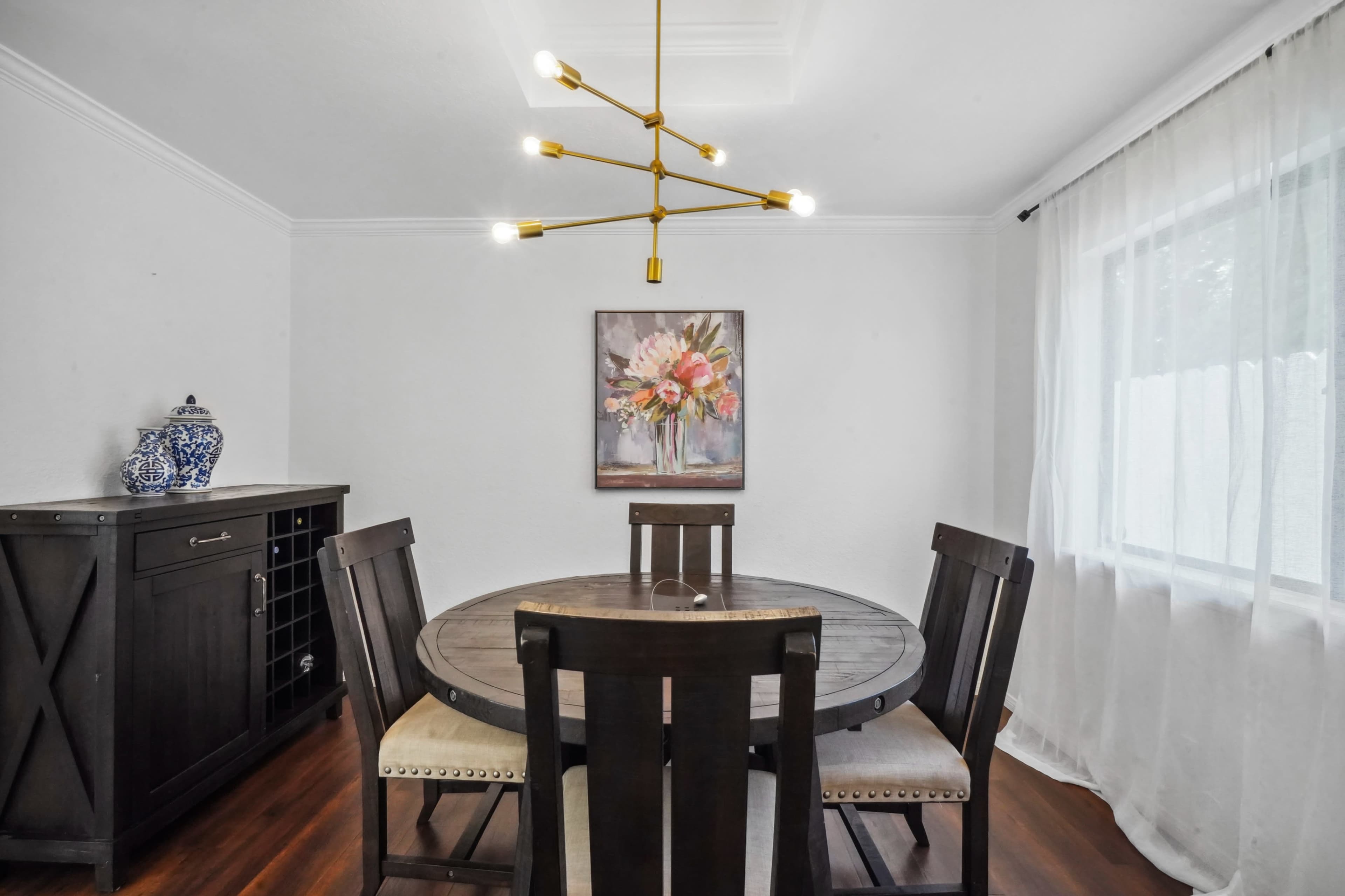A round dining table with four chairs is set in a brightly lit dining room featuring a floral painting on the wall and a side cabinet.