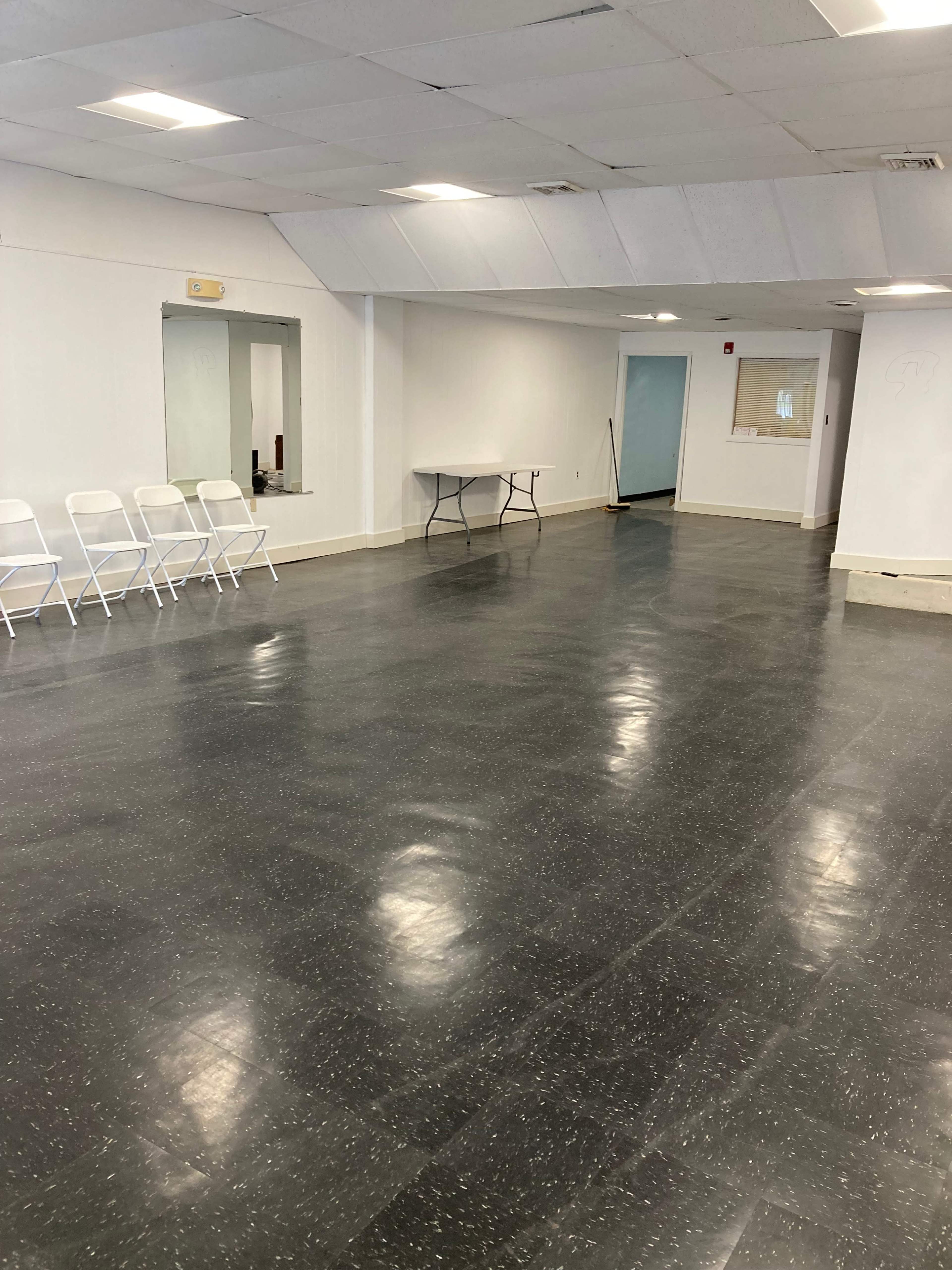 A large, empty interior space with a polished floor, a few white folding chairs lined up against one wall, and a table in the center.