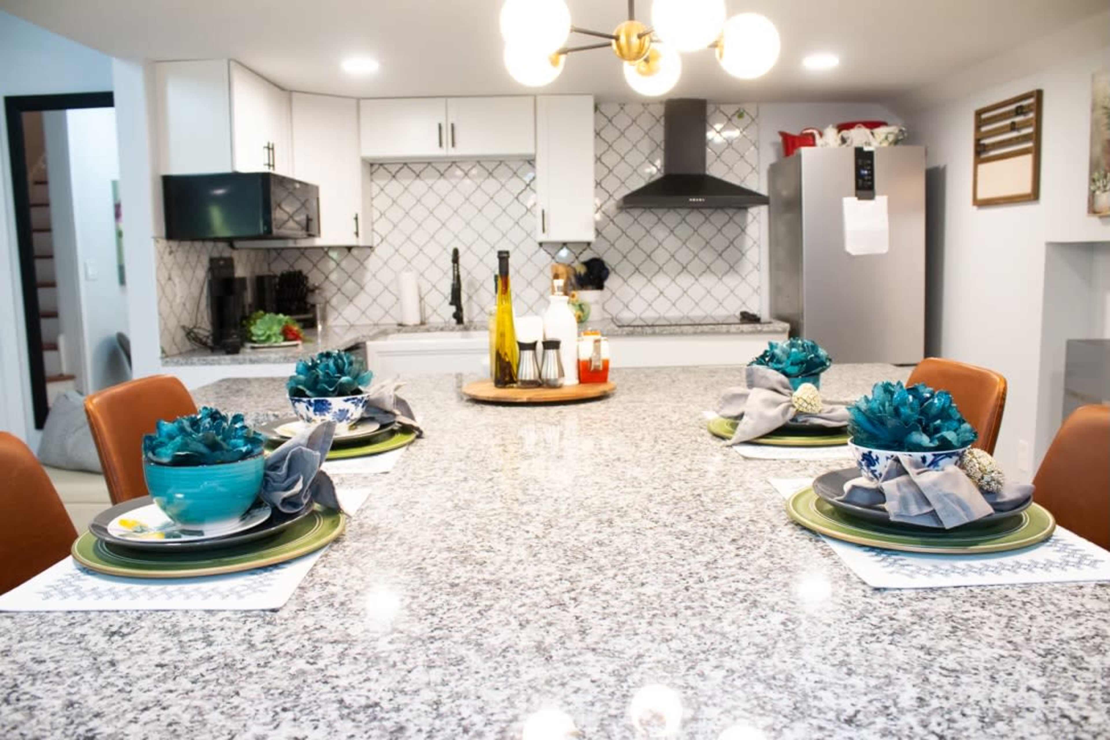 The image depicts a modern kitchen with a large granite island set for four, featuring teal tableware and a central decorative tray on a light gray countertop.