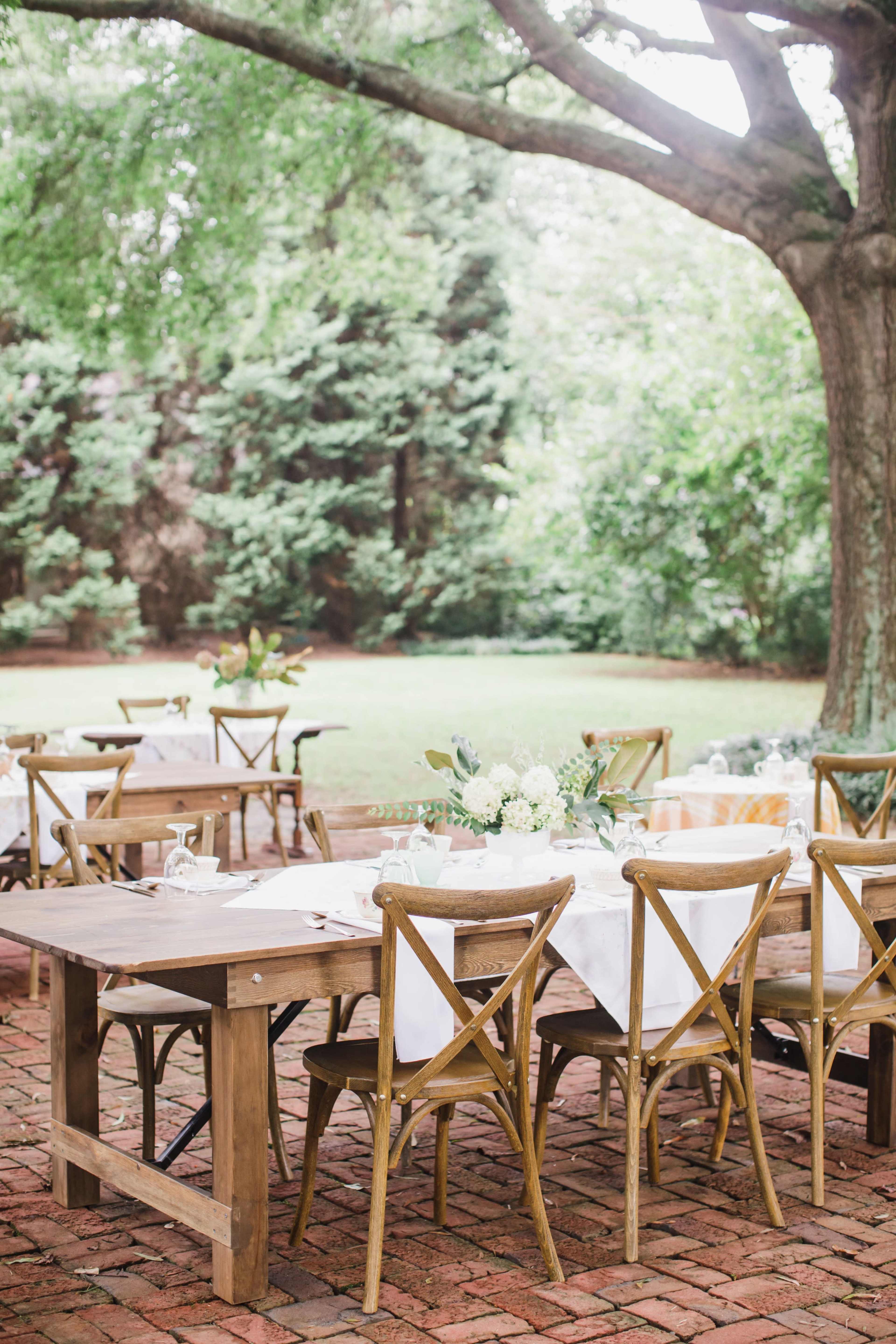 A rustic outdoor dining area features wooden tables and chairs set on a brick patio under a large tree, surrounded by greenery.