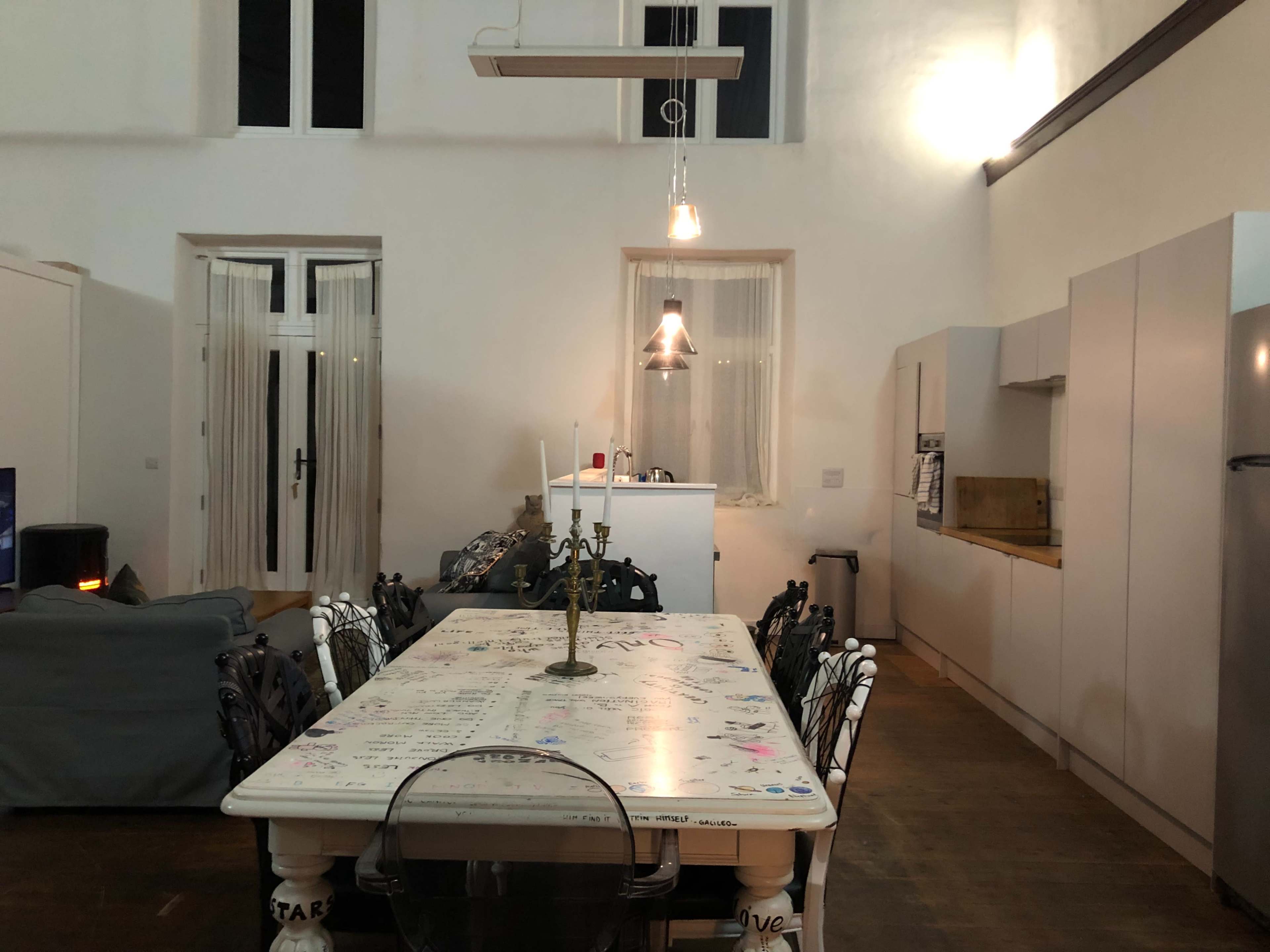 A long dining table with a brass candlestick is set in a spacious kitchen that features white cabinetry and a large window.