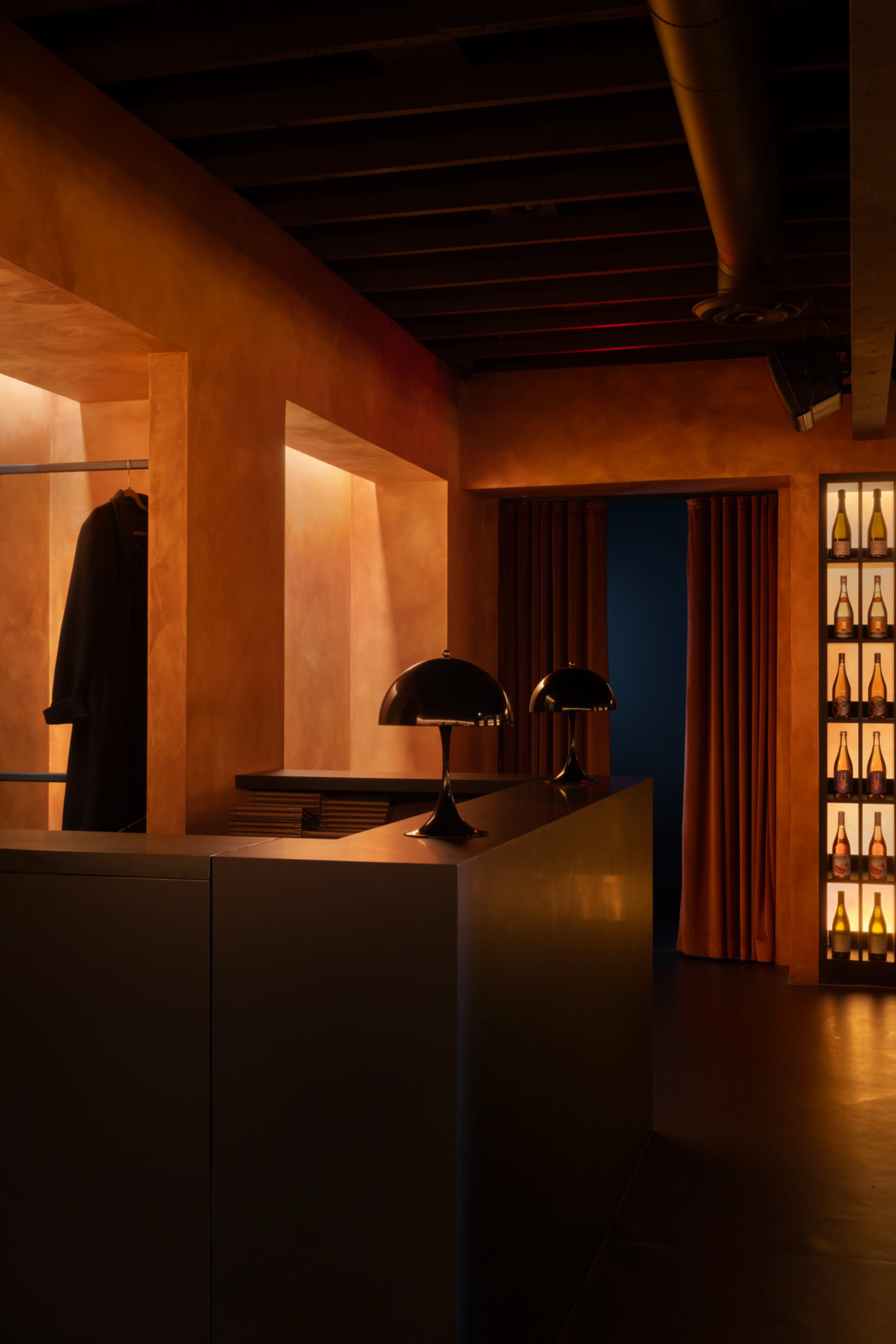 The image shows a warmly lit interior of a stylish bar or lounge with a sleek counter, a coat hanging on the wall, and shelves displaying bottles.