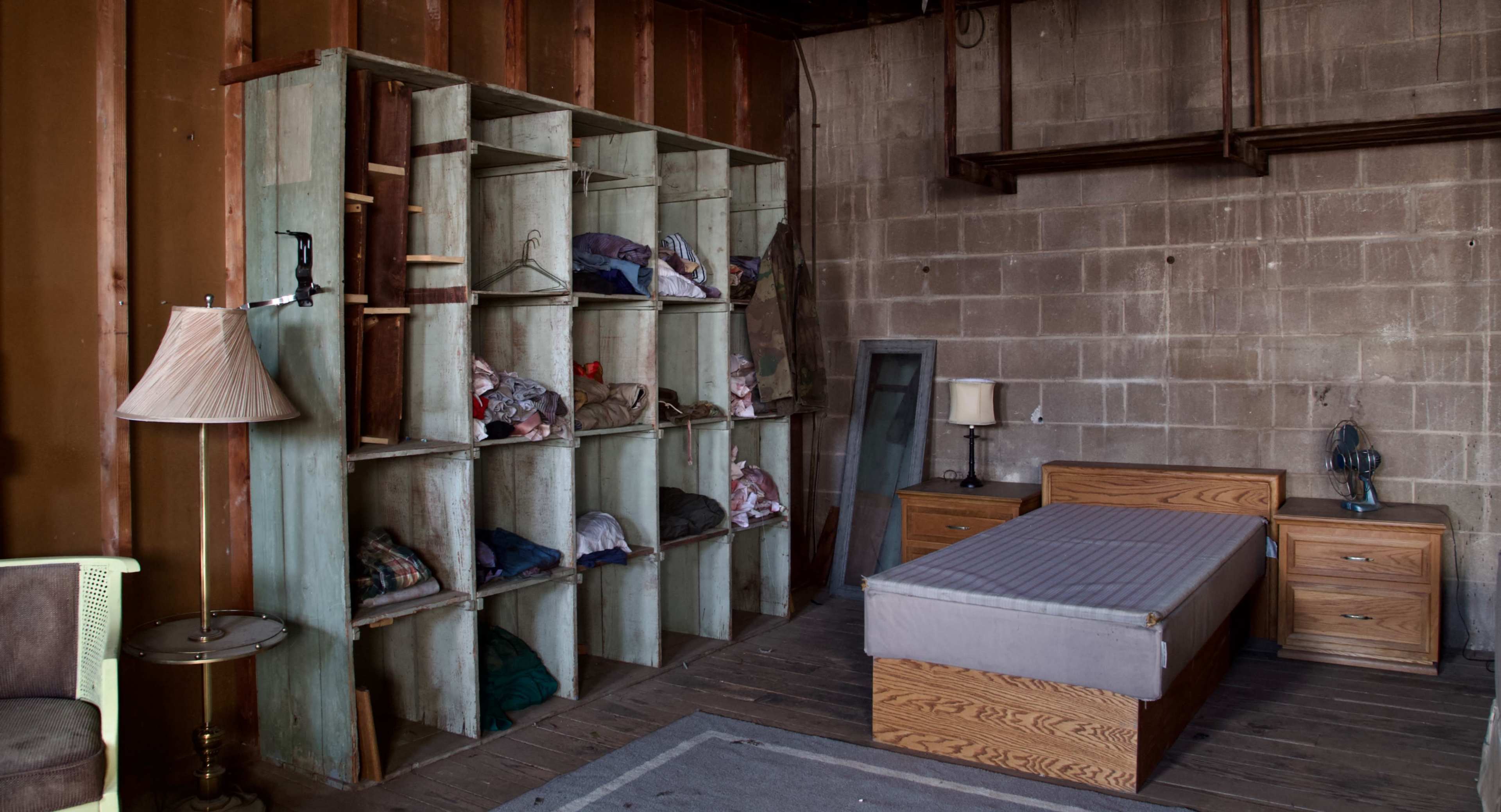 A sparse room with a single bed, wooden furniture, a lamp, and an open shelving unit filled with neatly arranged clothes.