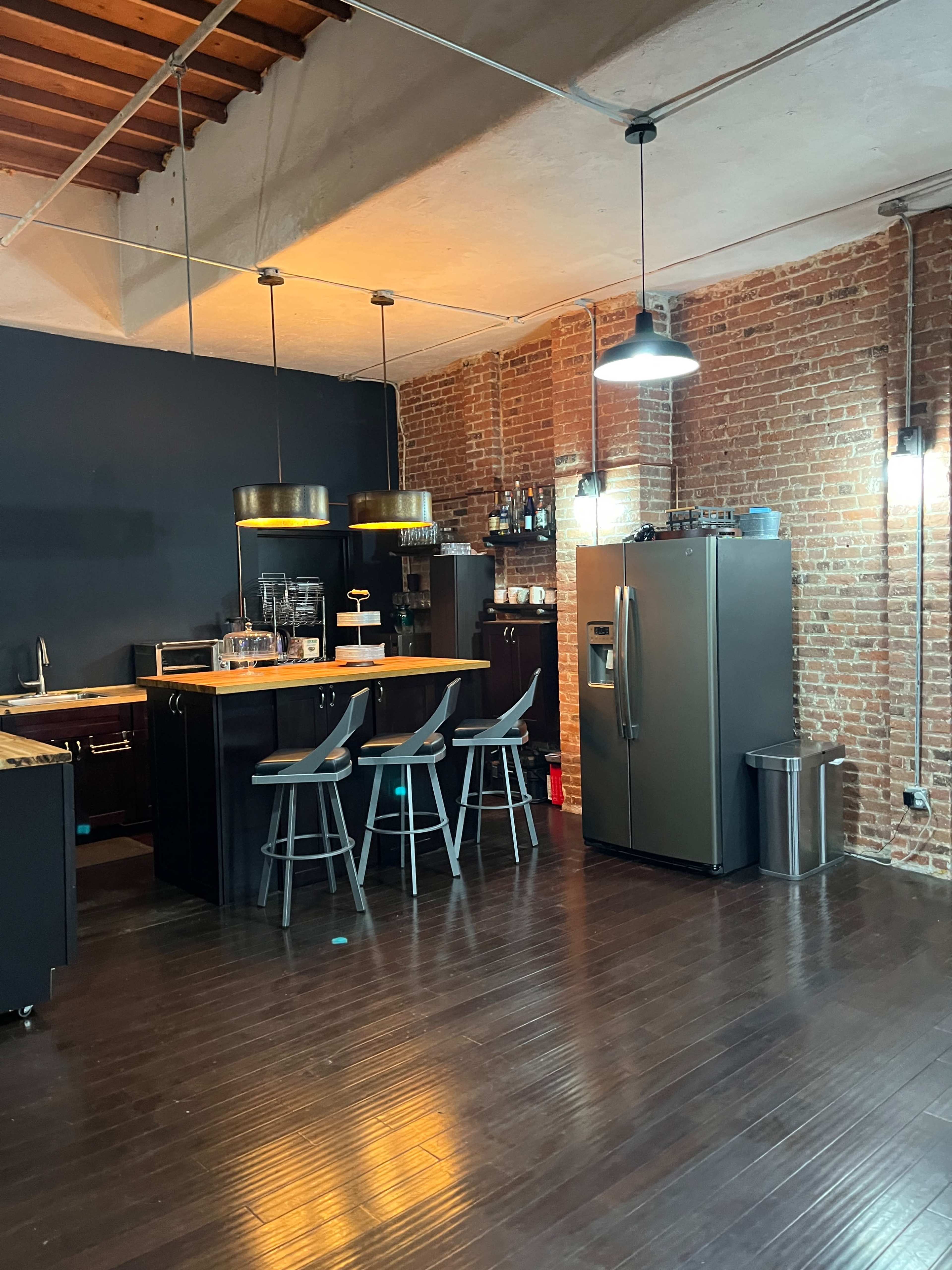 The image shows a modern kitchen with a bar counter, high stools, exposed brick walls, and stainless steel appliances.