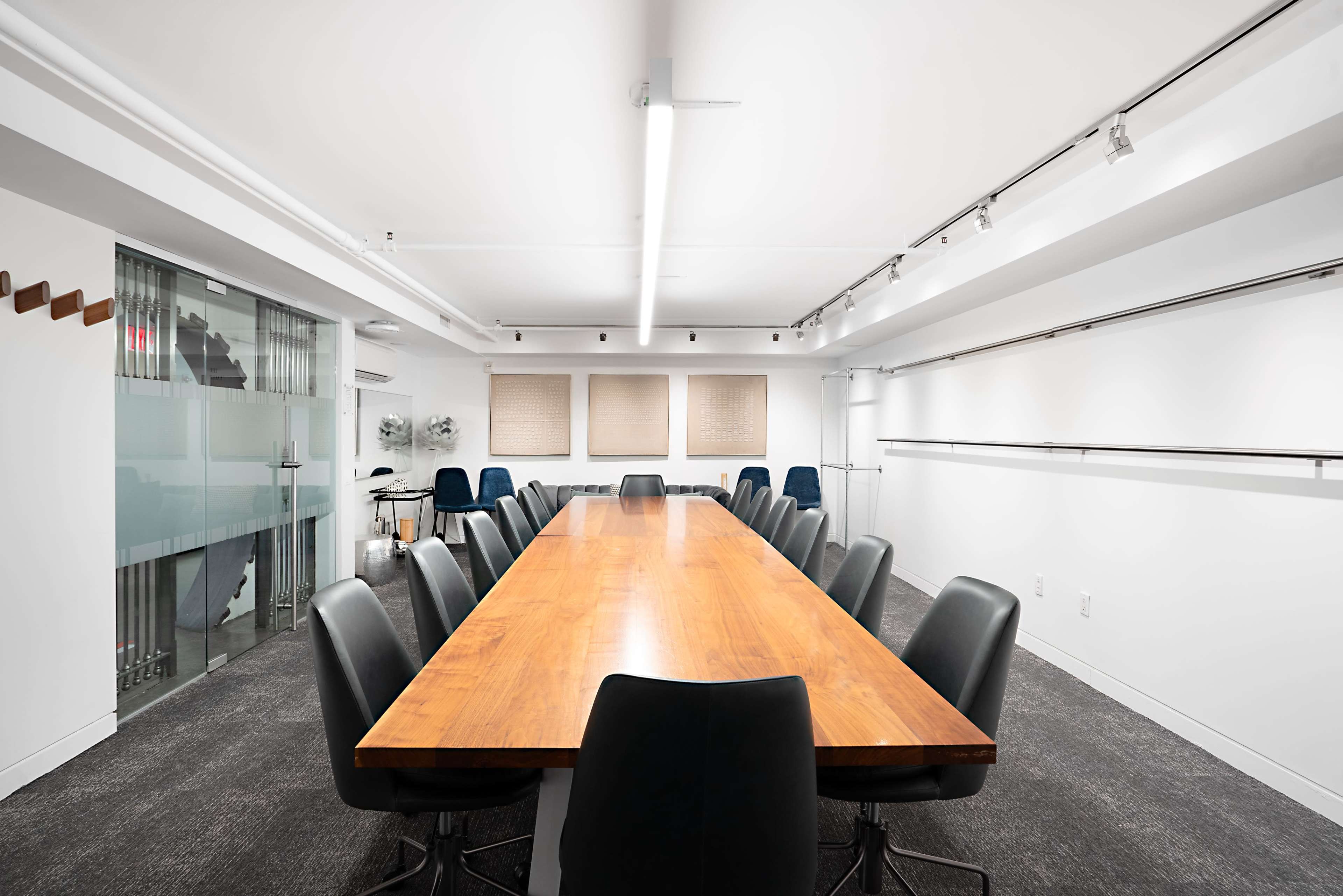 A long wooden conference table is surrounded by black chairs in a modern, well-lit meeting room with white walls and large windows.
