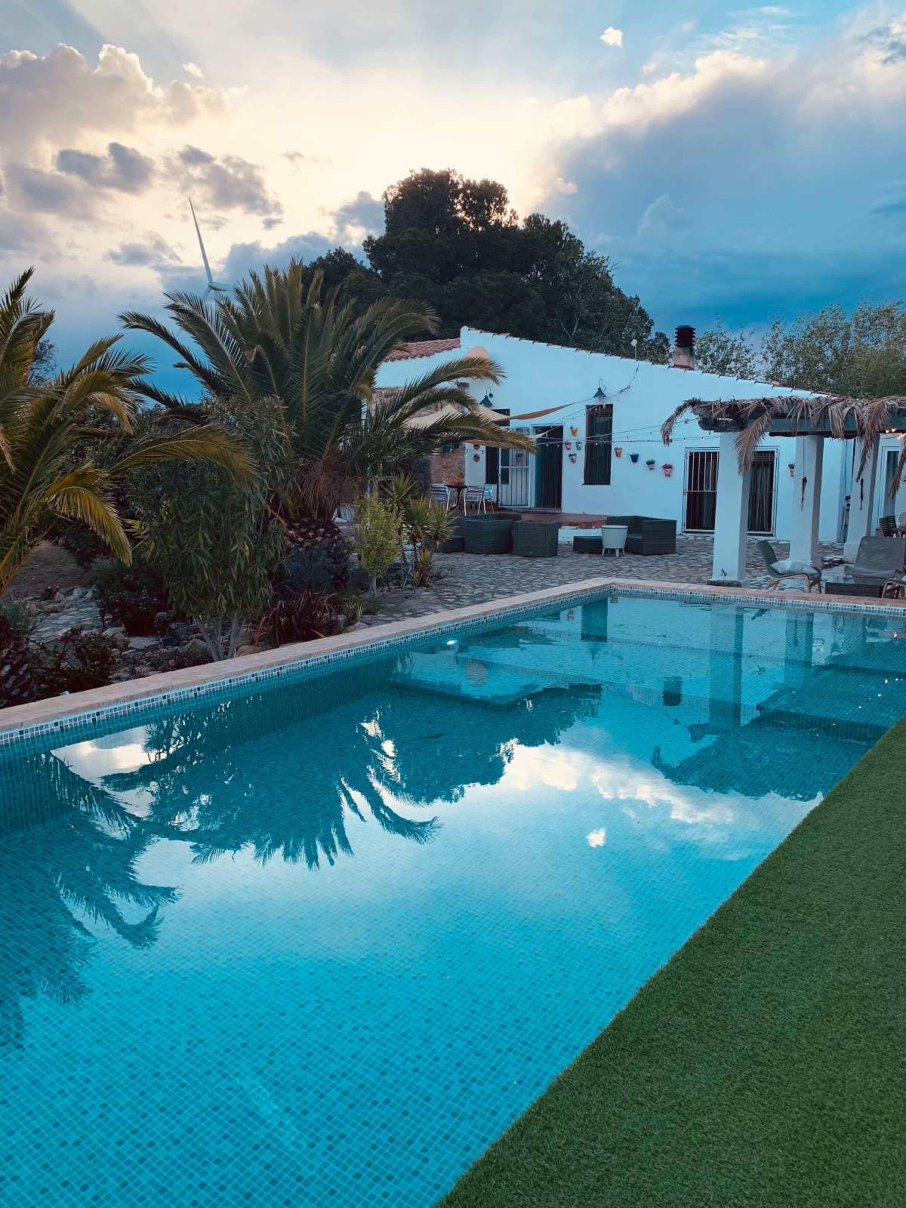 The image shows a tranquil swimming pool reflecting the sky and surrounding greenery in front of a white house.