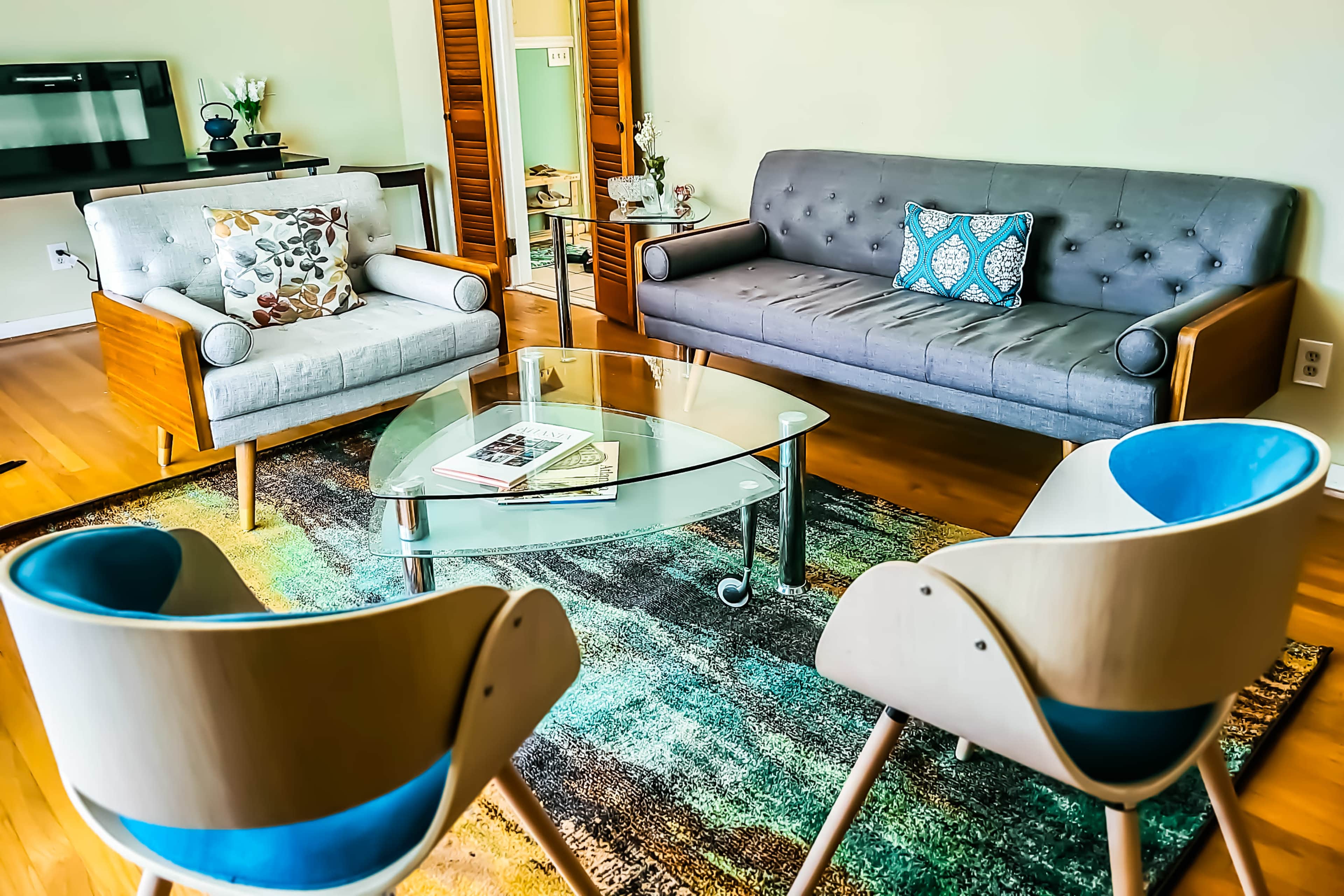 The image shows a modern living room with a gray sofa, two armchairs with blue and wooden accents, a glass coffee table, and a patterned rug on hardwood floors.