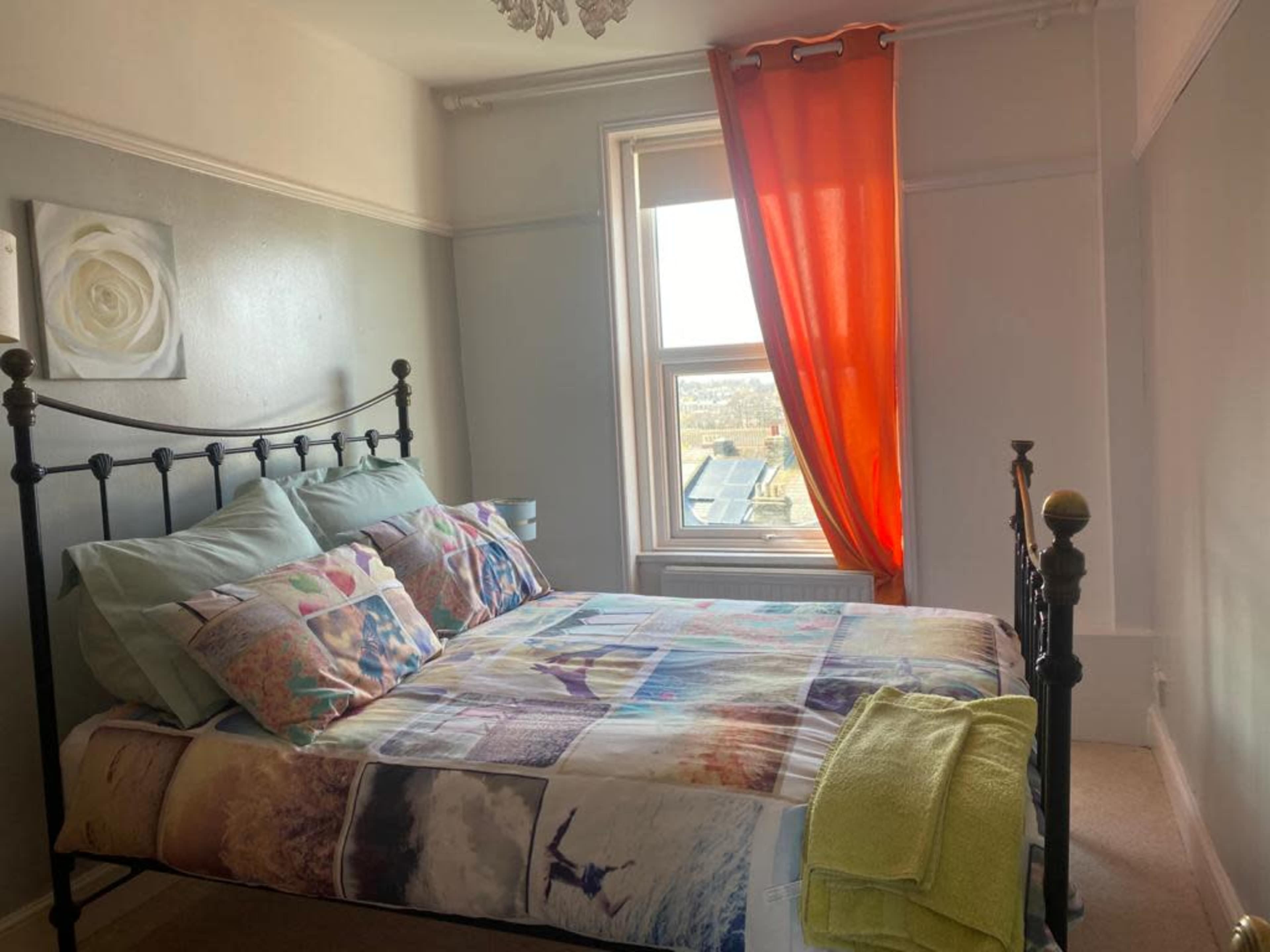 A bed with a multicolored quilt and several patterned pillows is positioned next to a window adorned with an orange curtain.