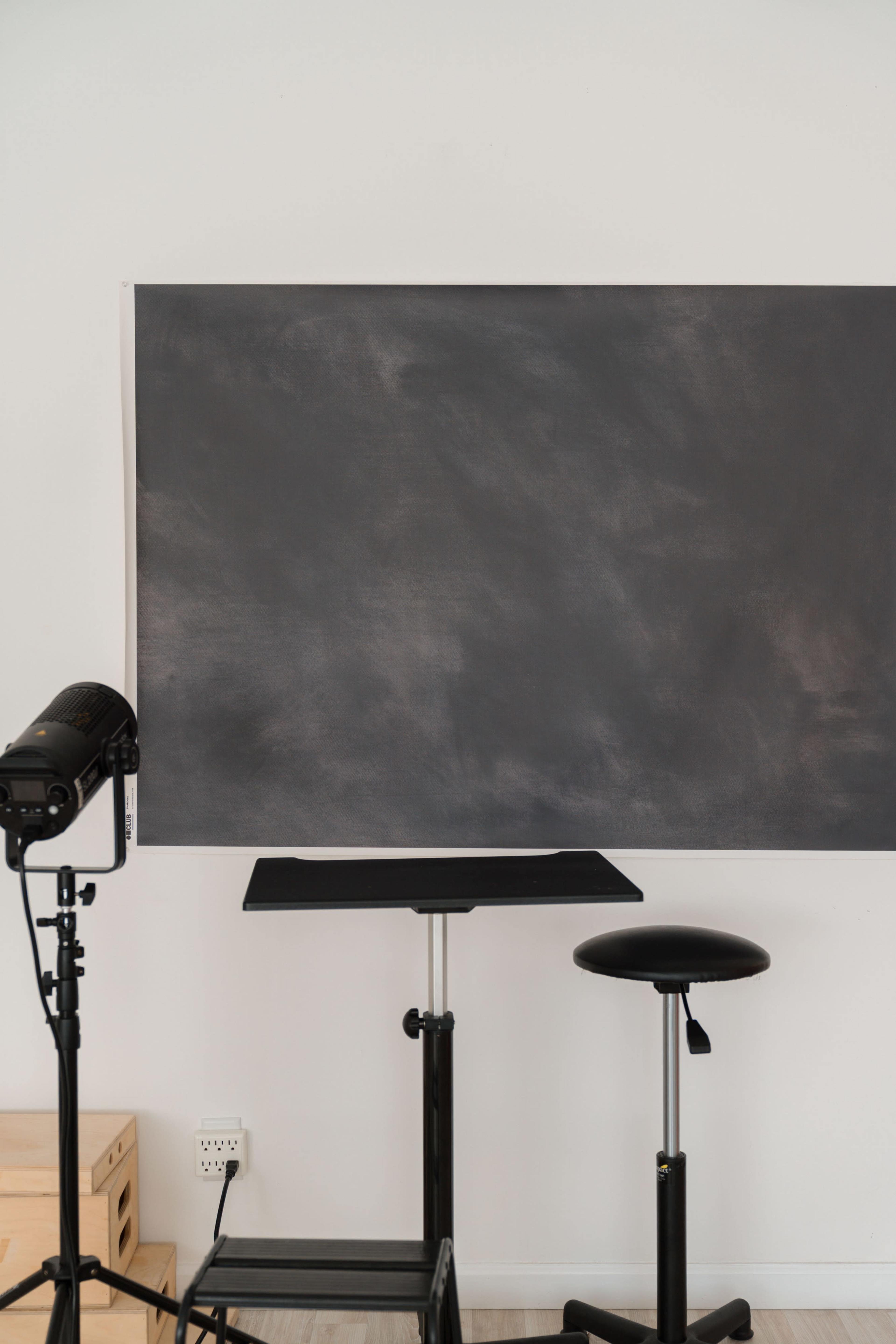 The image shows a minimalistic setup featuring a large blackboard, a small table, and a stool, alongside a spotlight on a tripod.