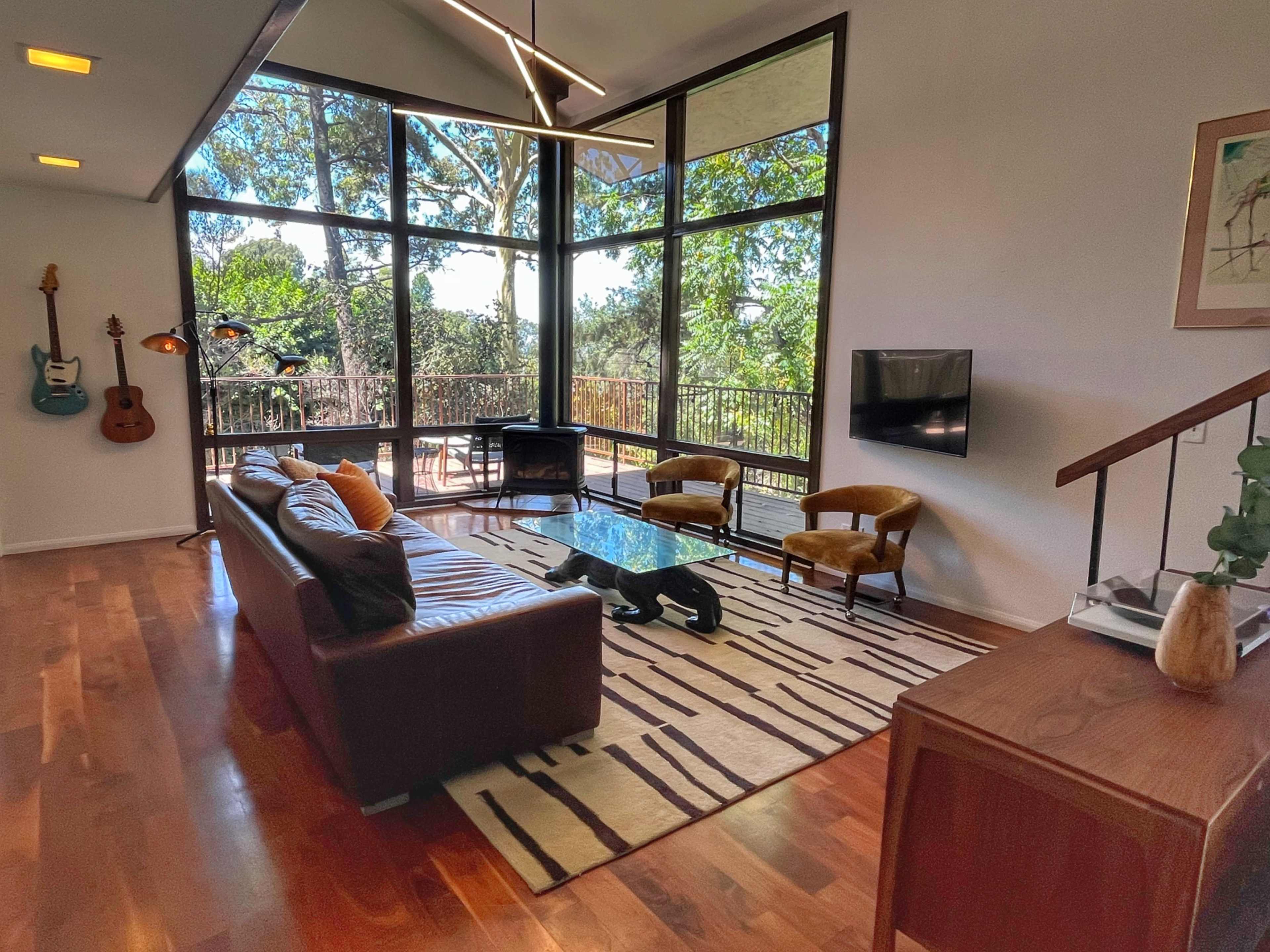 The interior of a modern living room features a leather sofa, two armchairs, a glass coffee table, and large windows that overlook a wooded outdoor area.