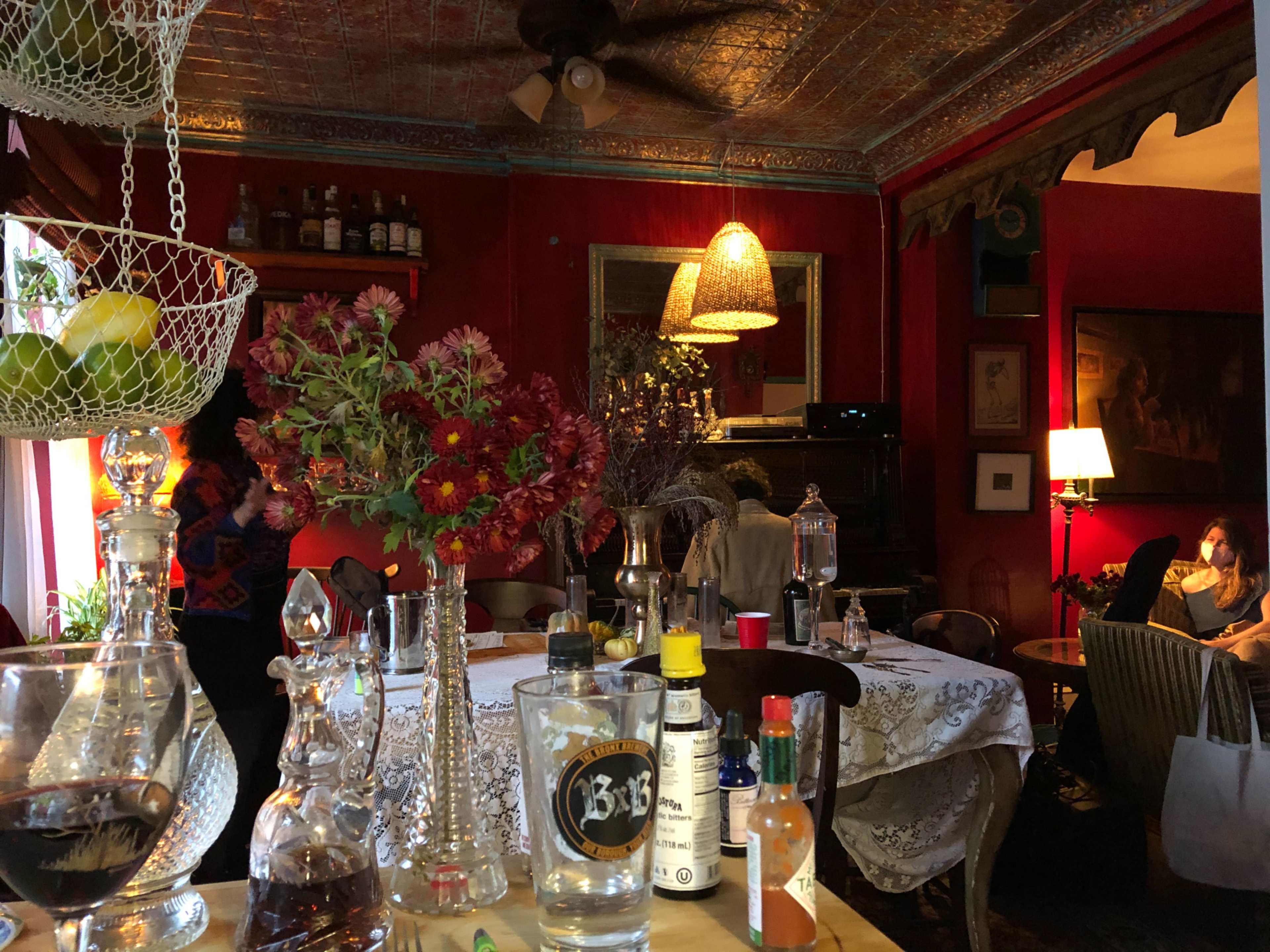 The image shows a cozy restaurant interior with a table set for dining, flowers in glass vases, and various bottles on display against a backdrop of red walls and vintage decor.
