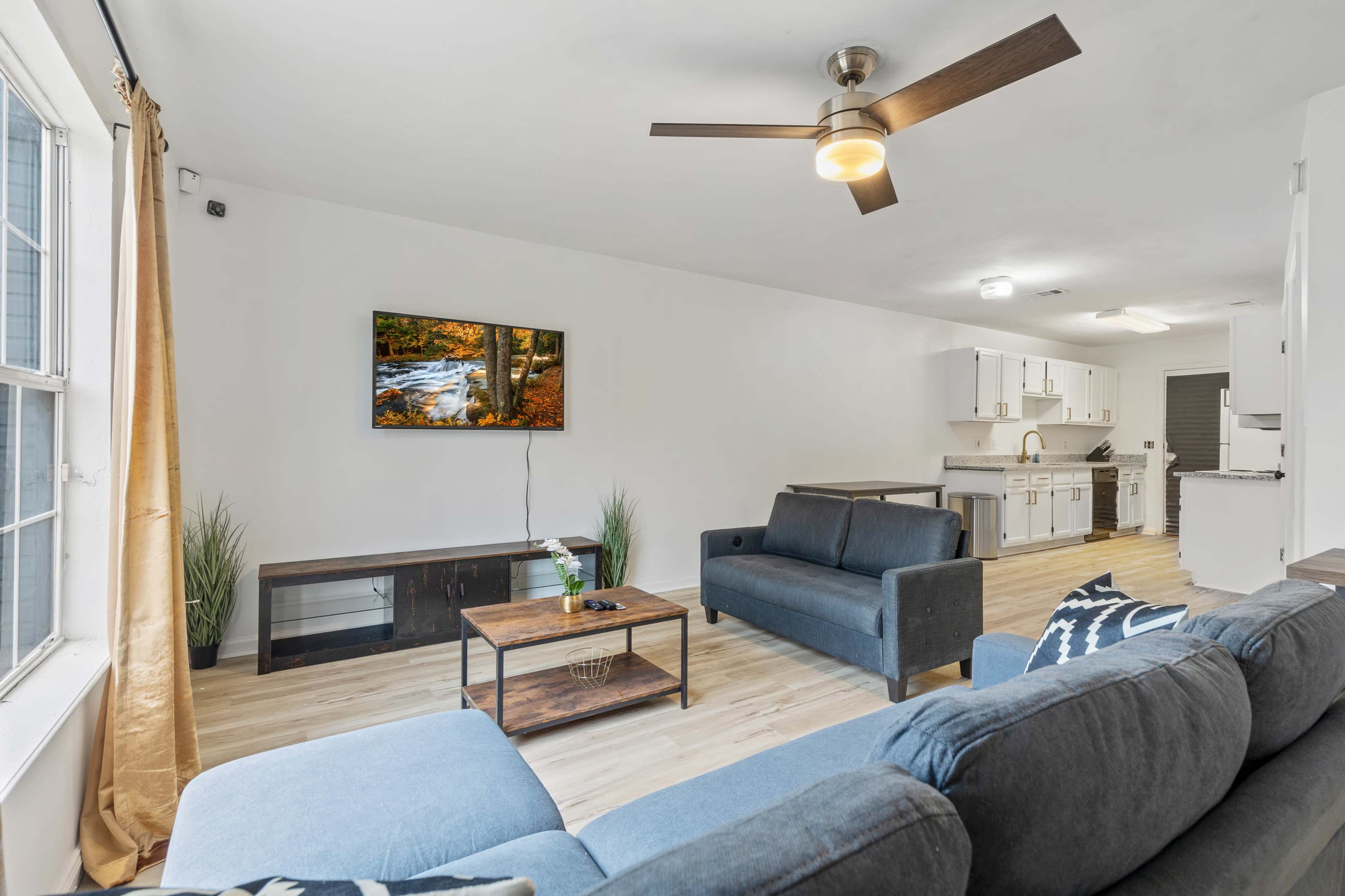 The living room features a television mounted on the wall, two sofas, a coffee table, and a kitchen visible in the background.
