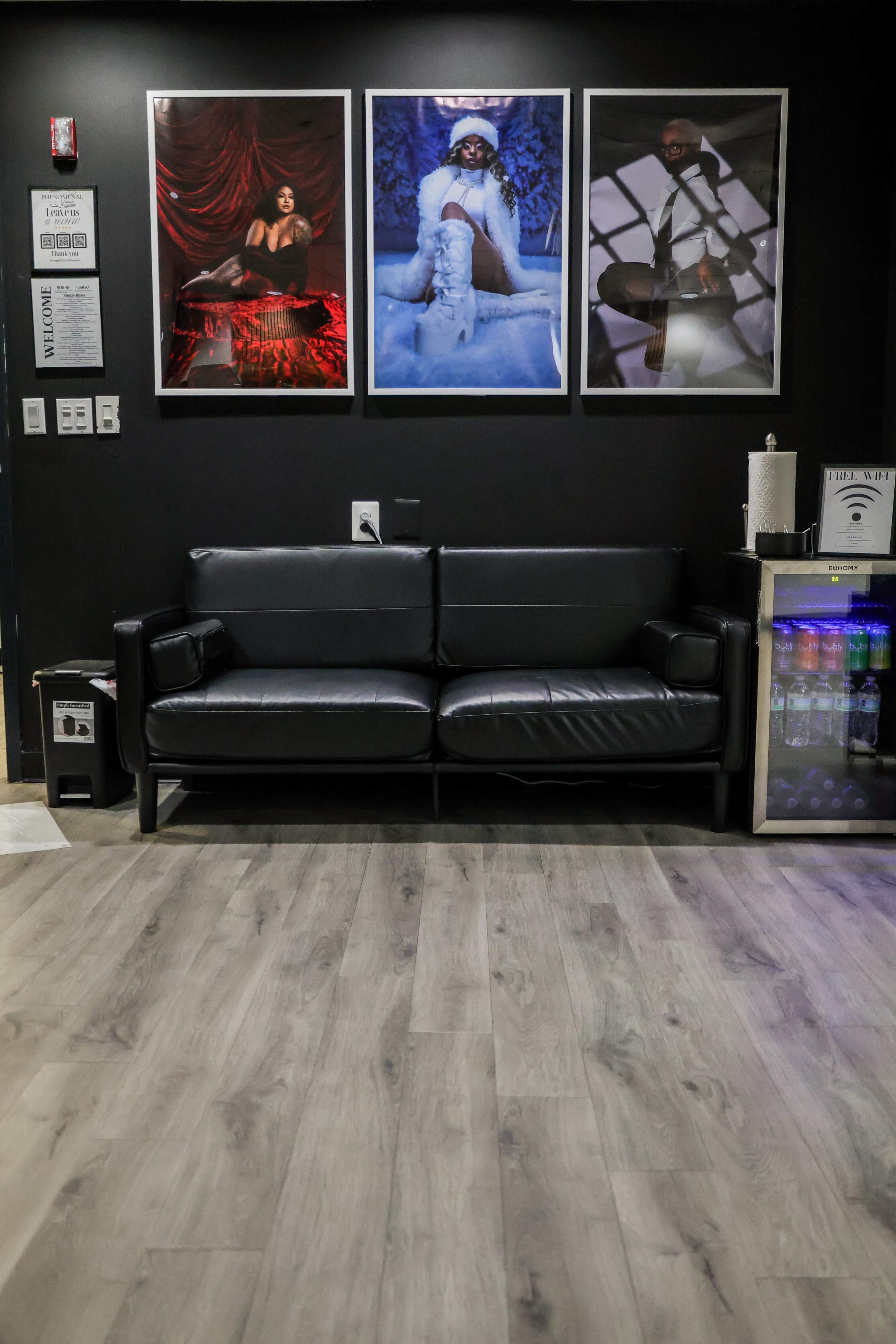 The image shows a black leather sofa in front of three framed photographs on a dark wall, with a small beverage station to the right.