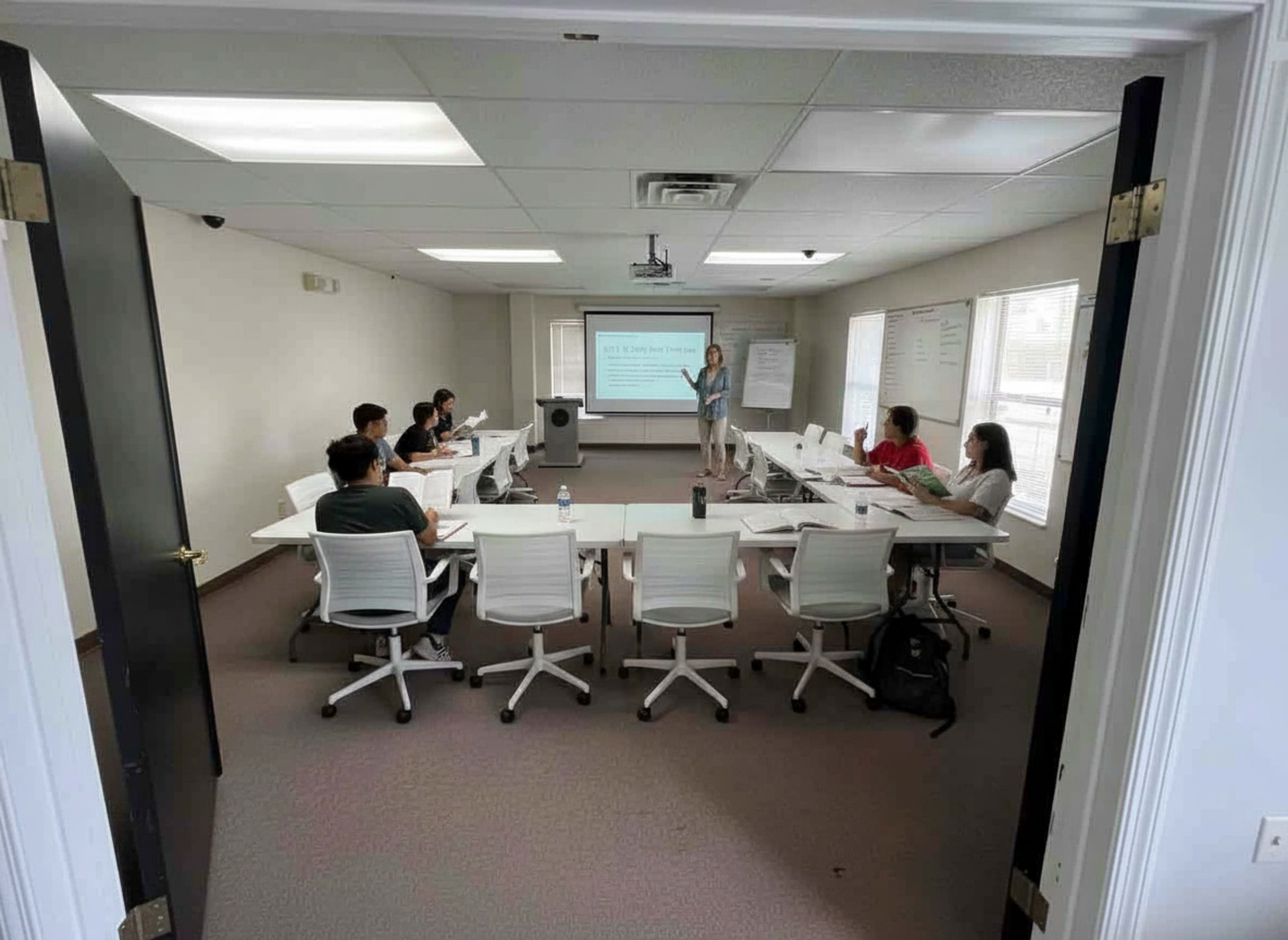 A group of people sits around a large table in a classroom while a presenter stands in front, speaking to them.