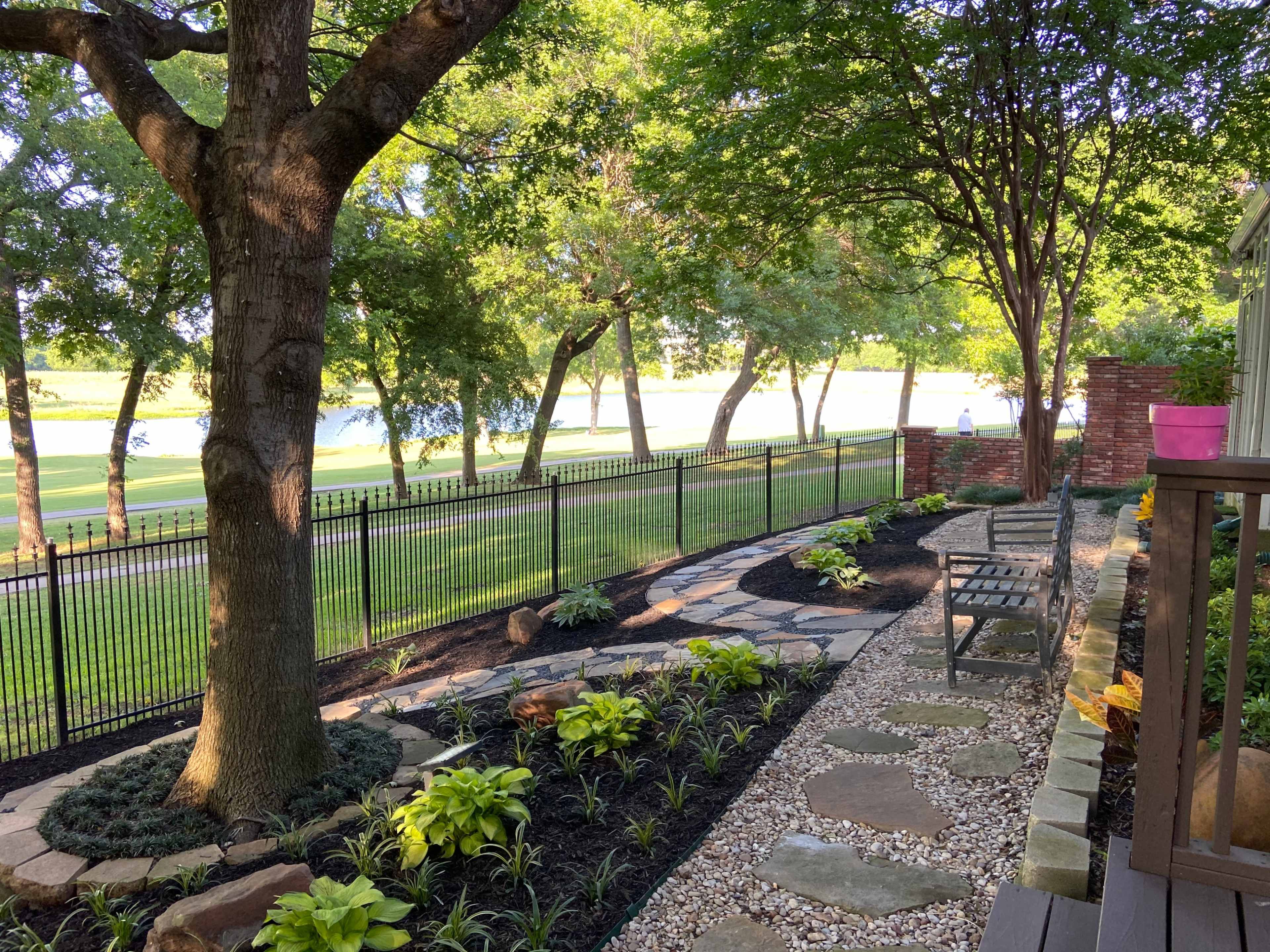 A landscaped garden with stone pathways, plants, and a view of a fenced area next to a body of water.