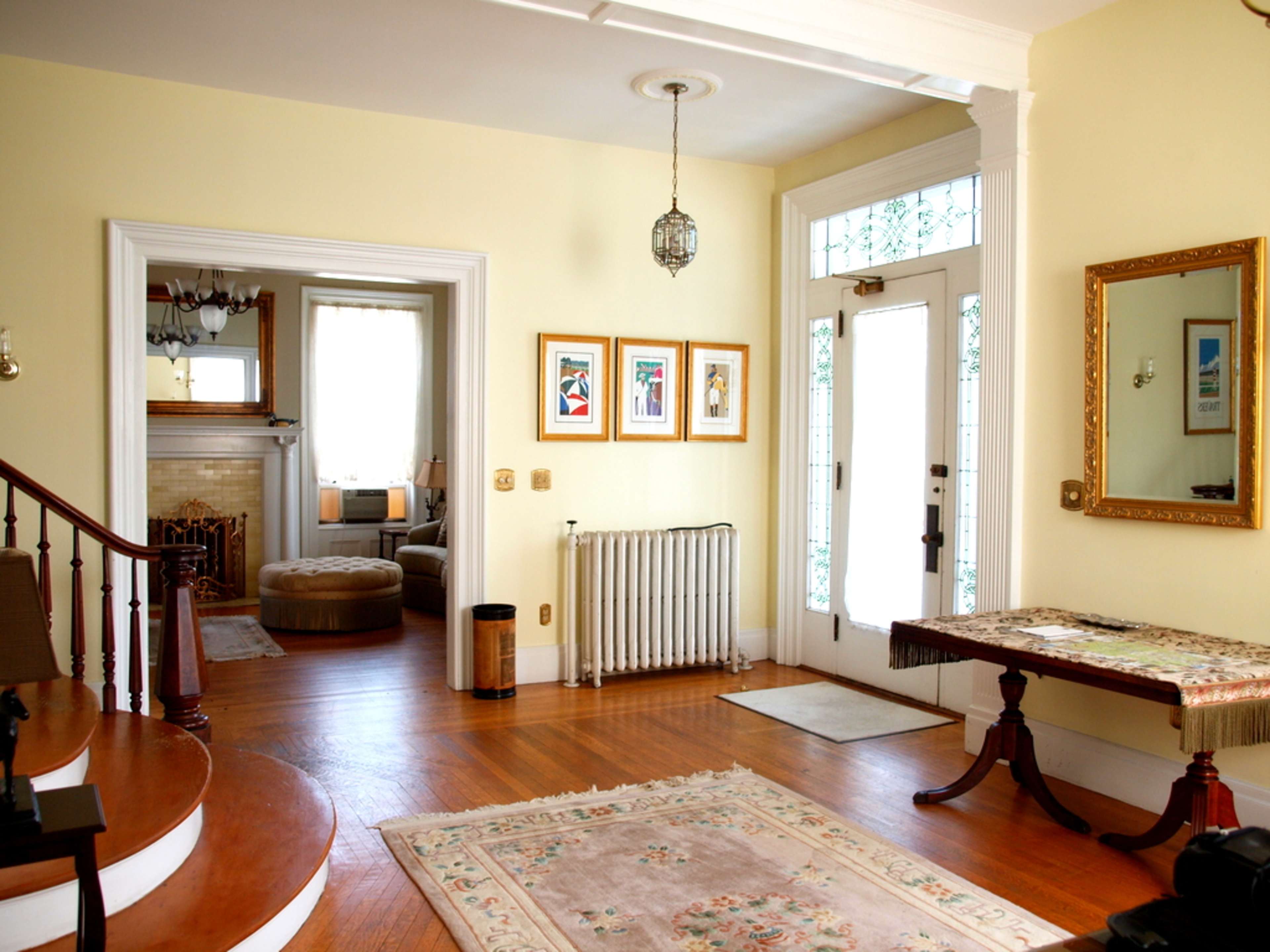 The image shows a bright entryway with a staircase, framed art on the wall, a radiator, and a door leading to an adjacent room.