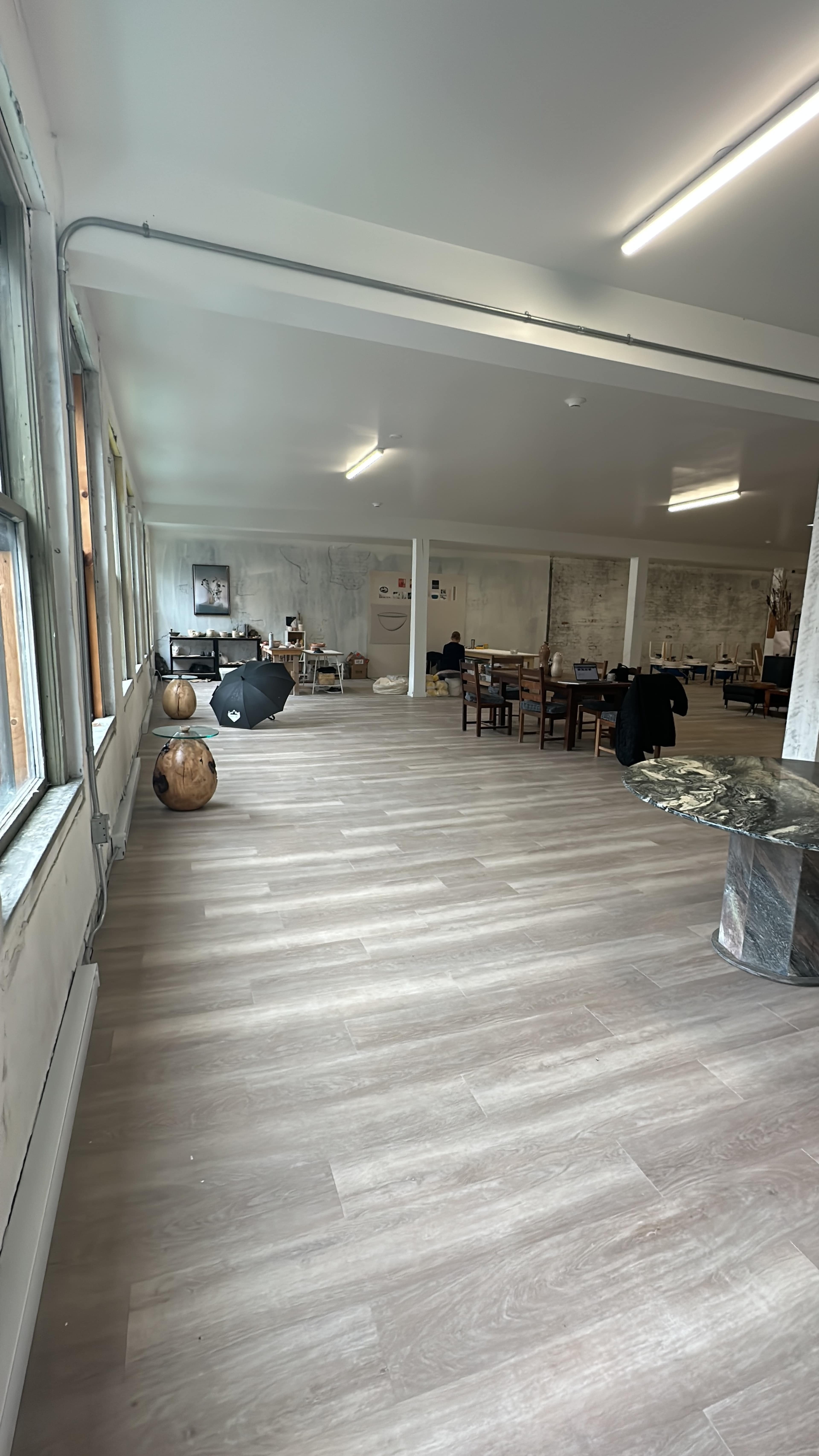 A spacious room with wooden flooring, large windows, a dining area with tables and chairs, and a few decorative objects.