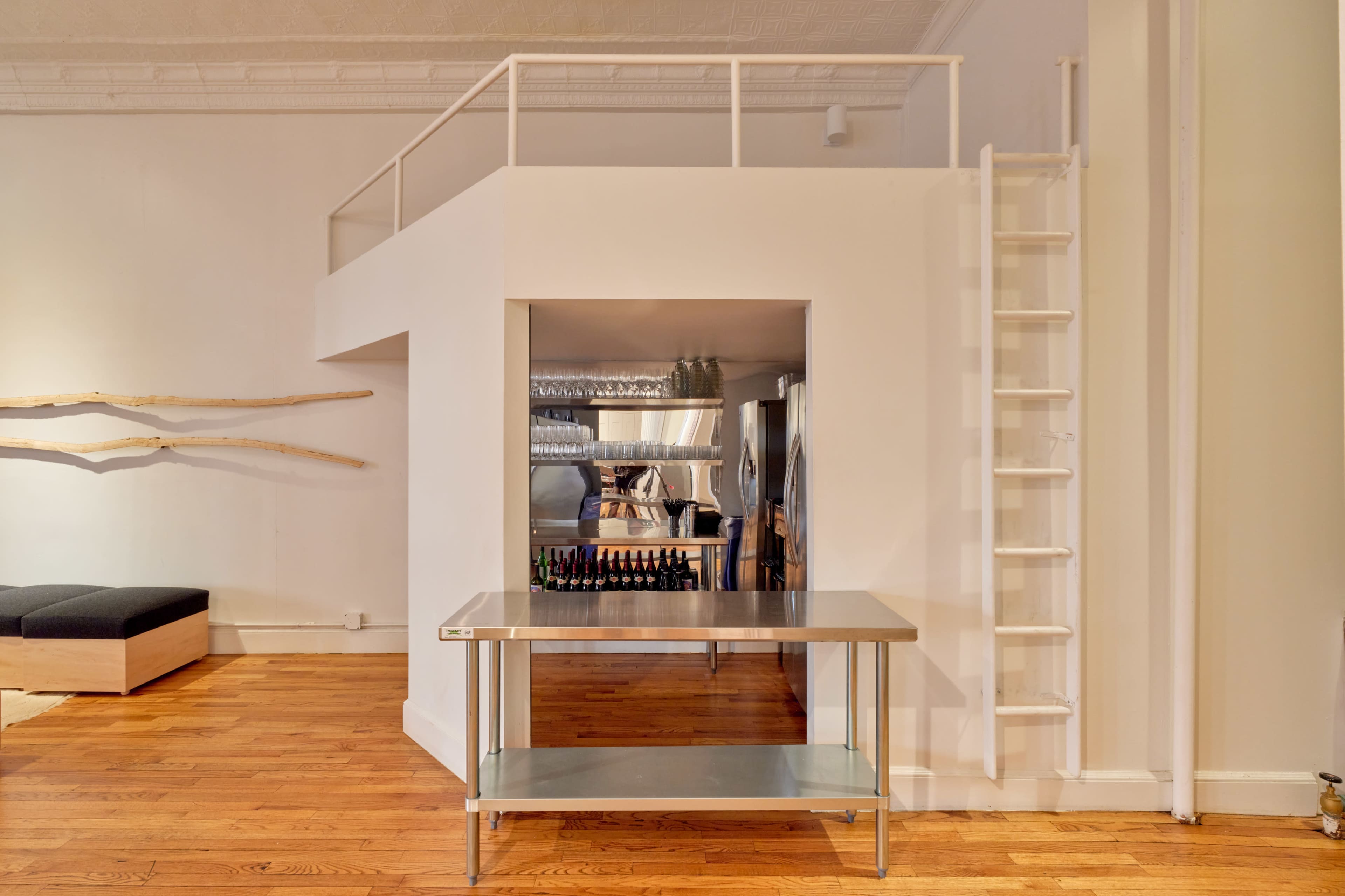 The image shows a minimalist interior space featuring a light-colored wall with a raised platform, a metal table in the foreground, and a ladder leading to the platform.