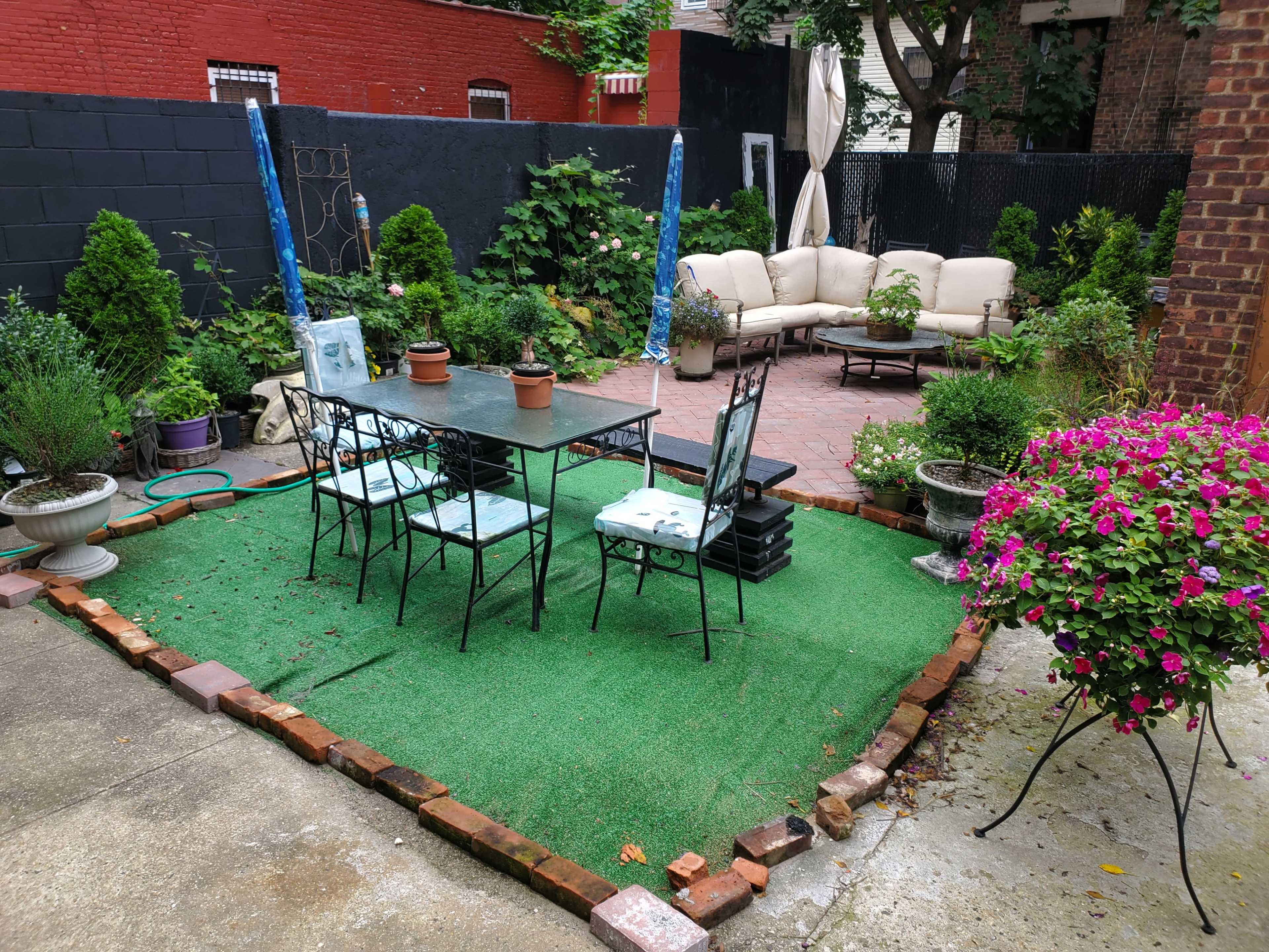 A backyard area with a green artificial grass surface, a dining table surrounded by chairs, and a seating arrangement with a sofa in the background, all framed by various plants and flowers.