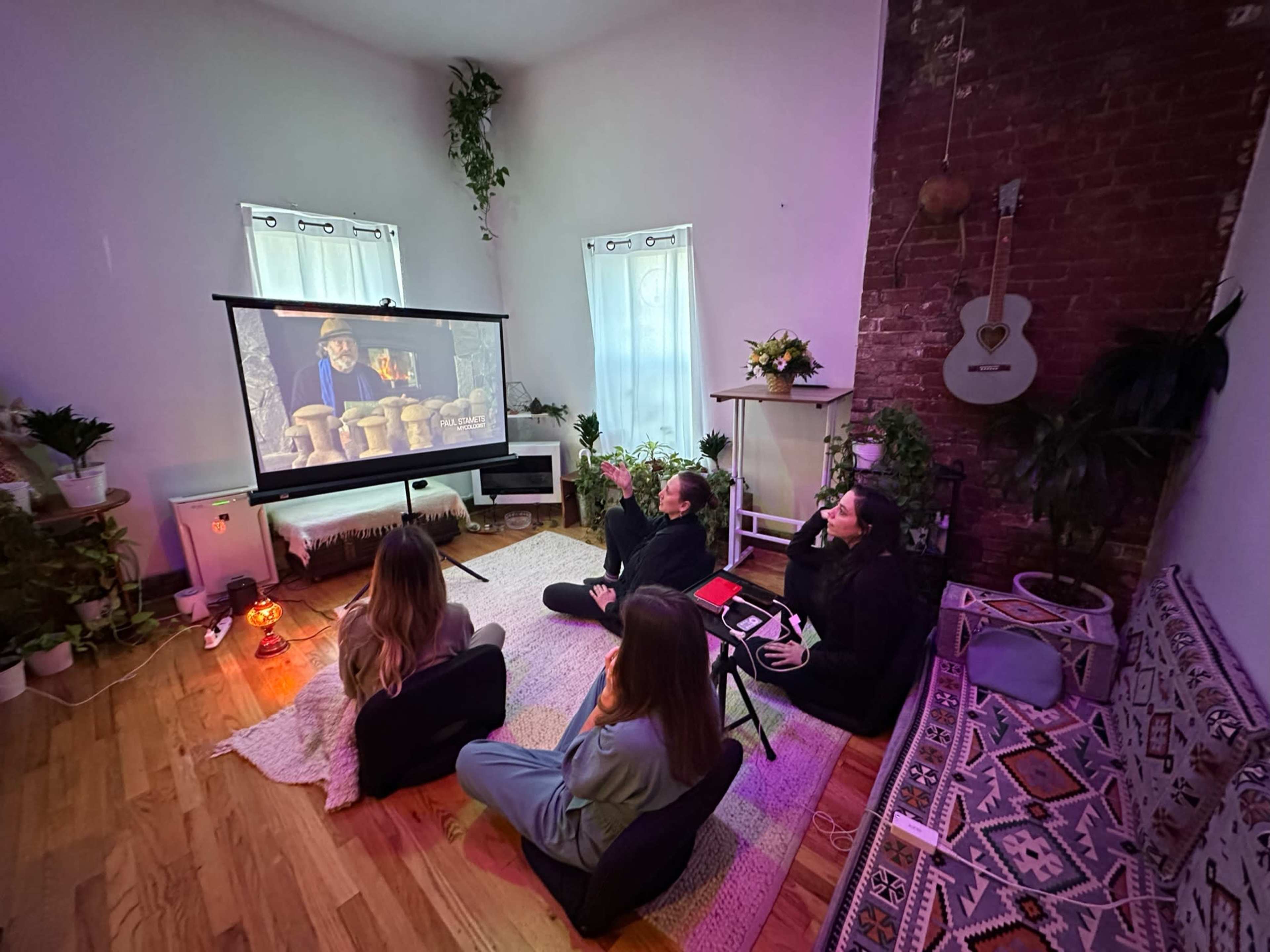 Four people sit on floor cushions in a cozy room, watching a movie projected on a screen.