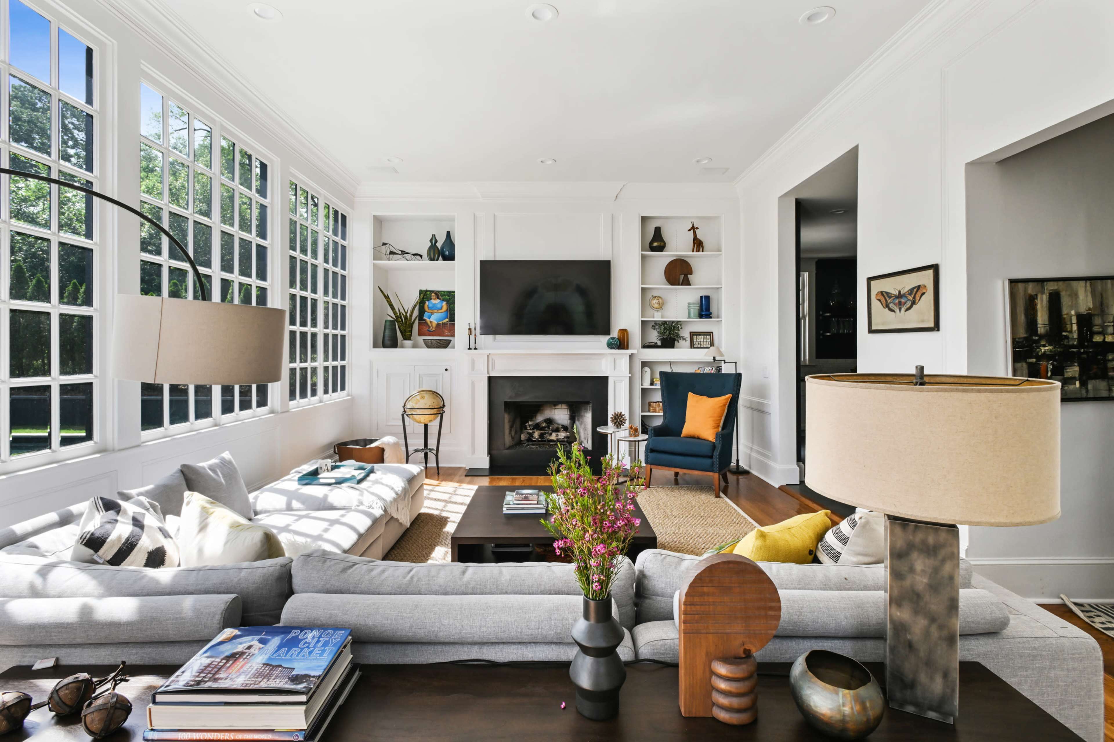 A bright living room features a large sectional sofa, a coffee table with decorative items, and a fireplace with a television above it, alongside windows letting in natural light.